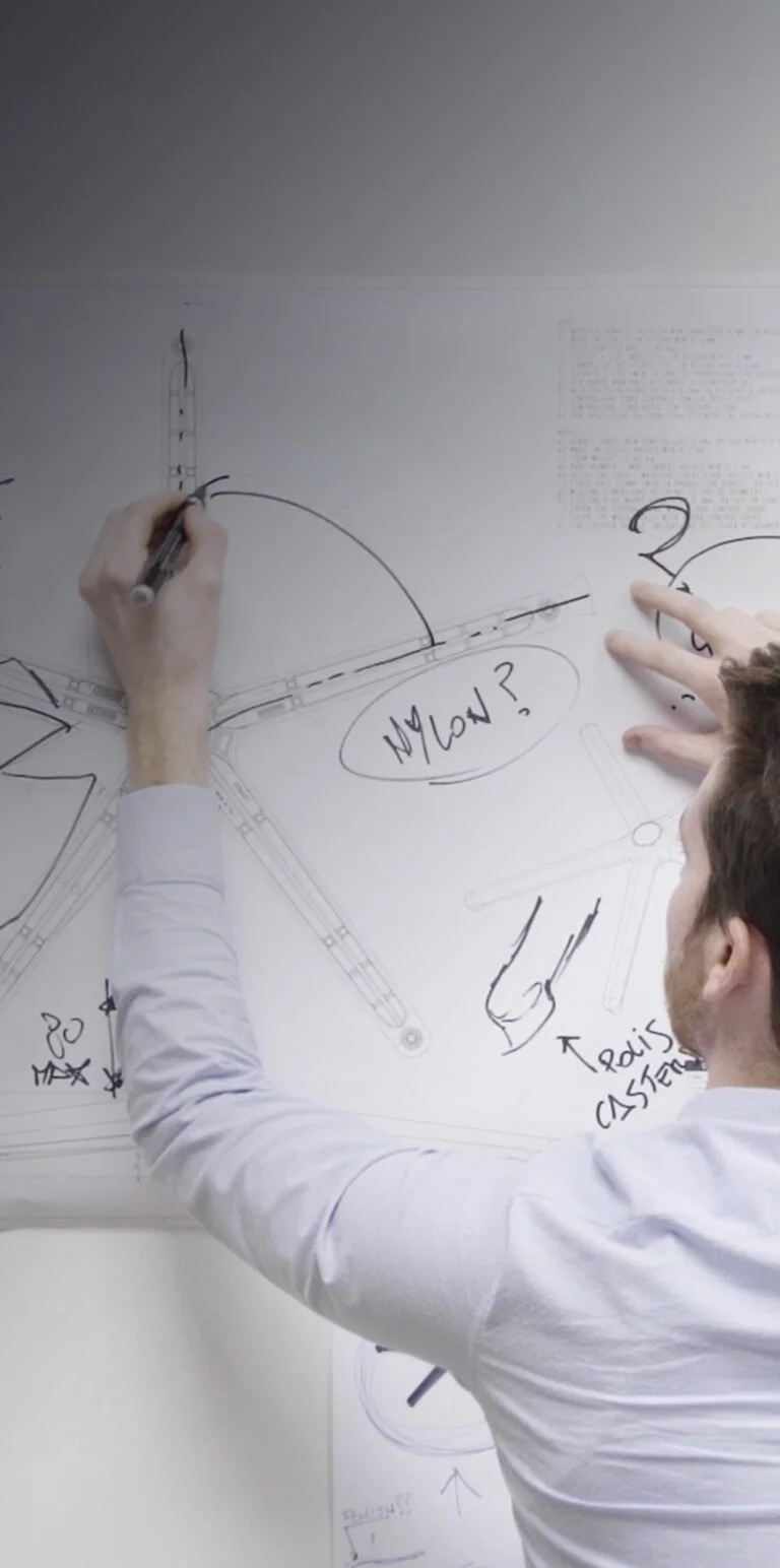 Person som ritar tekniska scheman på ett vitt papper, med text och pilar, i ett arbetsrum.