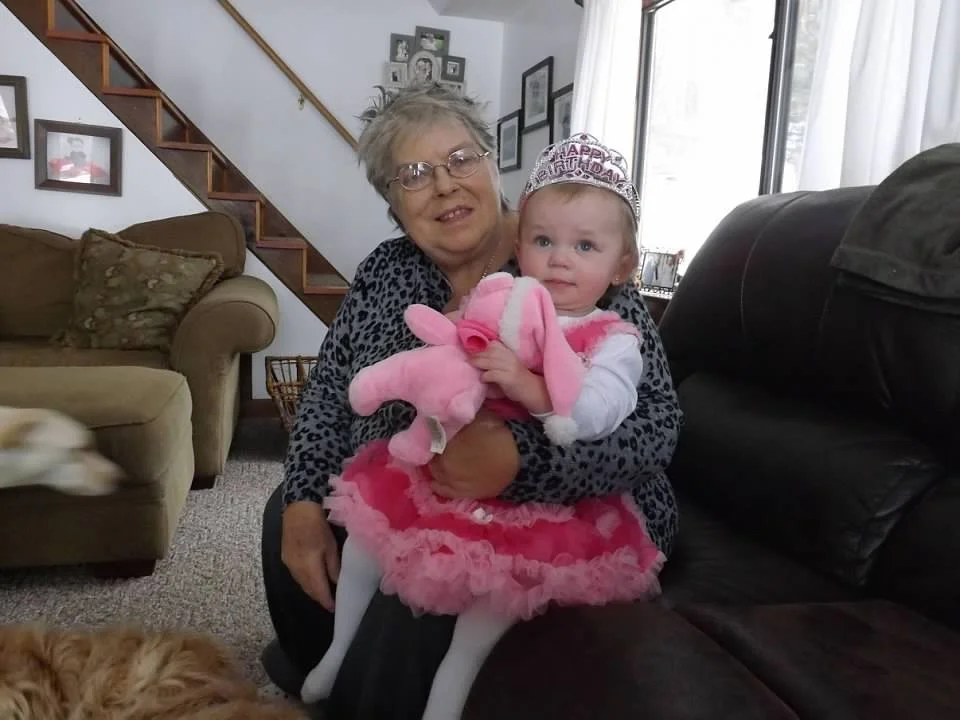 An elderly woman holding a young girl dressed in pink at a birthday celebration in a living room.