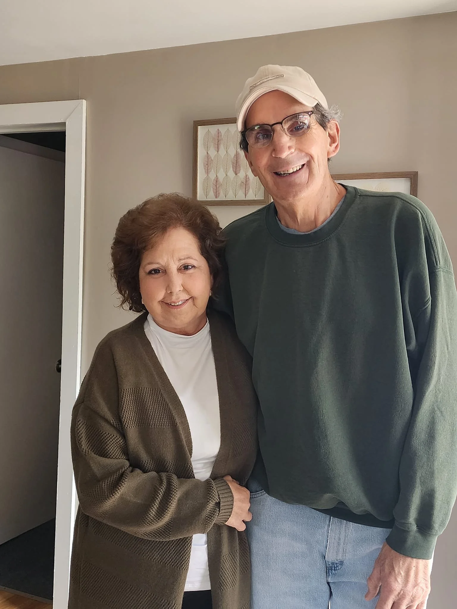 A middle-aged woman with brown hair wearing a white top and brown cardigan, standing next to a tall man with glasses, wearing a beige baseball cap, green sweatshirt, and light blue jeans, smiling at the camera inside a home.