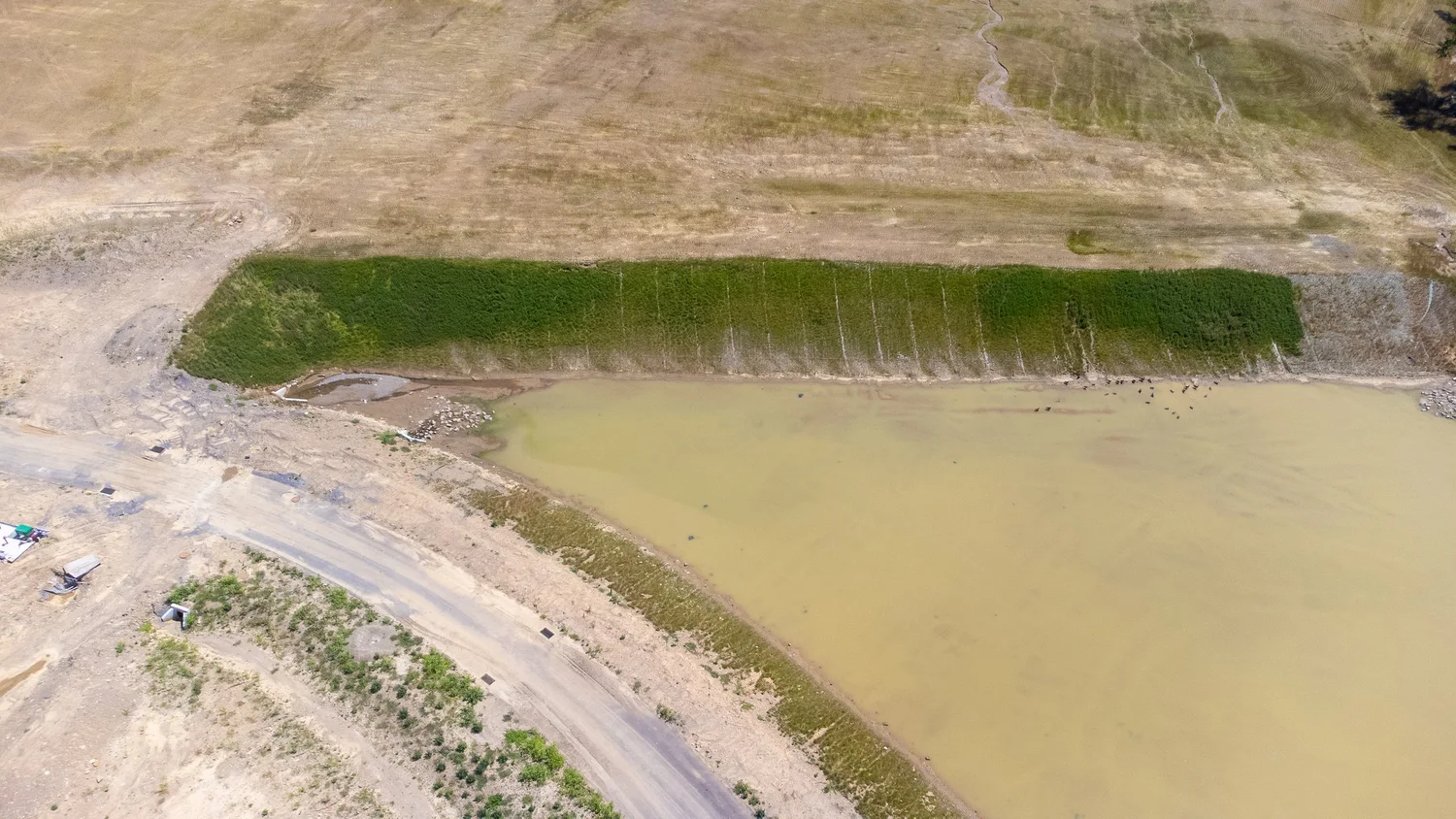 Aerial view of a construction site next to a body of water, with a large green area and dirt paths.