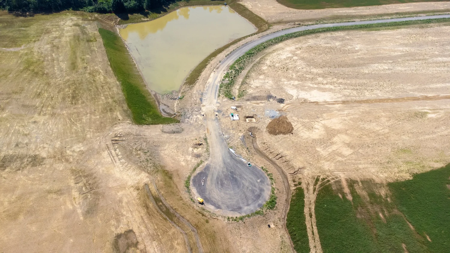Aerial view of a construction site with a circular driveway, dirt roads, and a pond, surrounded by fields and farmland.