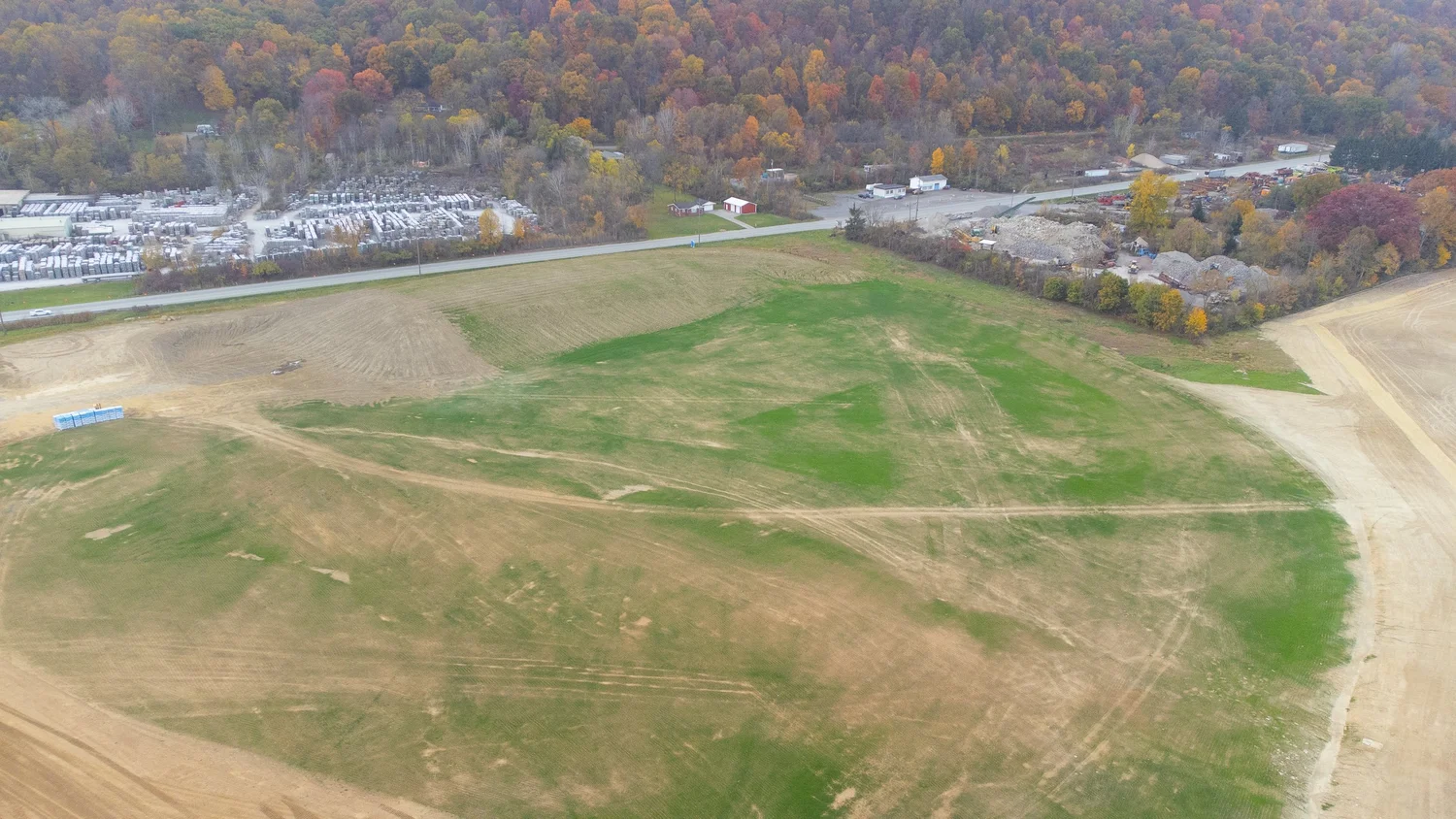 Aerial view of a landscape with a large open field, a paved road, and a forested area with colorful autumn trees.
