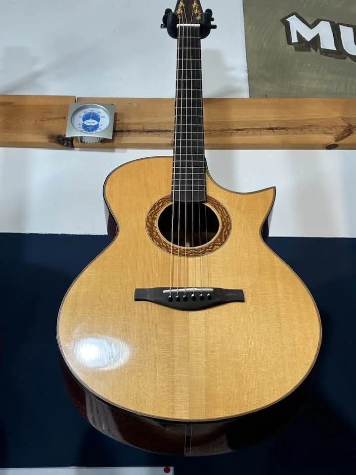 Handcrafted guitar by Tom Roeger. Luthier based in the midwest, available beyond. Tenor Ukulele. E model guitar. An acoustic guitar with a unique body shape and dark fretboard, resting on a surface with a poster and a wall clock in the background.