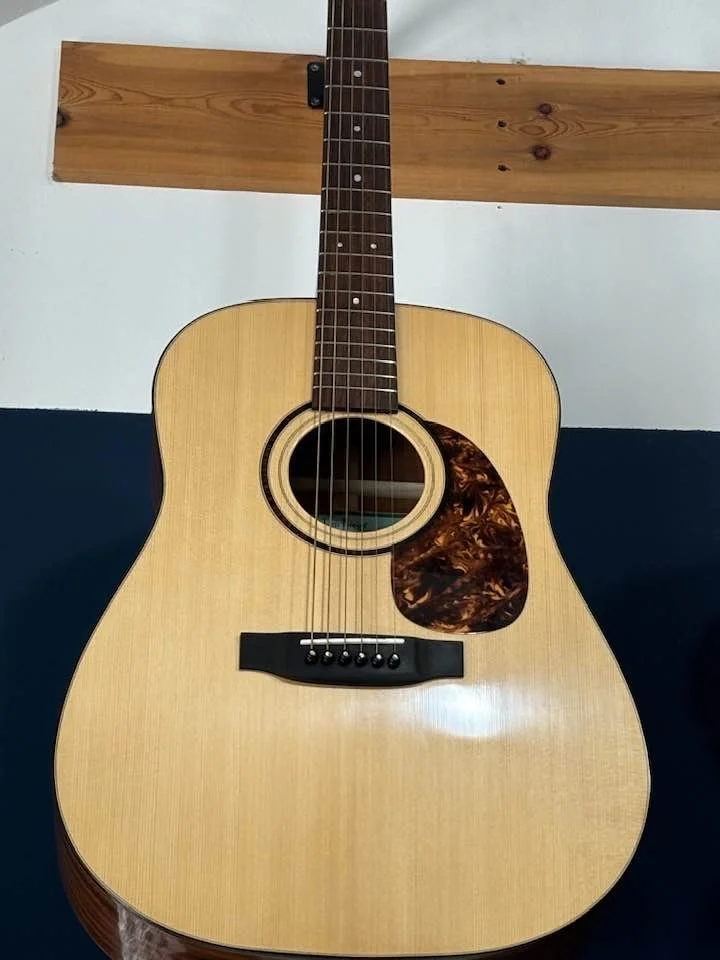 Handcrafted guitar by Tom Roeger. Luthier based in the midwest, available beyond. Tenor UkuleleAn acoustic guitar with a light wood body, dark fretboard, and a tortoiseshell pickguard, resting against a wall, with a wooden crossbar in the background.