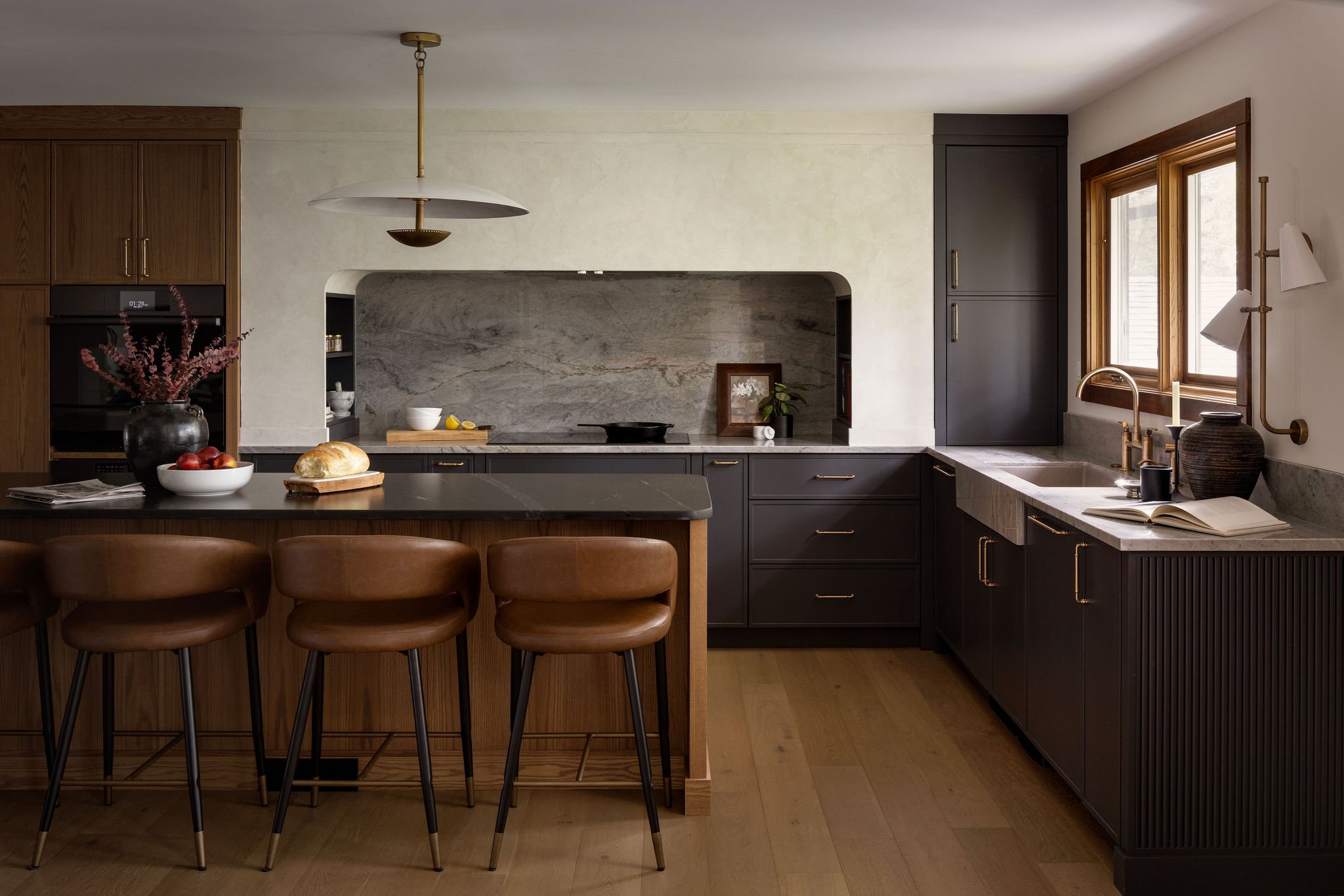 A modern kitchen with dark cabinets, a marble backsplash, a kitchen island with brown leather stools, a window with wooden trim, and various decor including a vase with pink flowers and a loaf of bread.