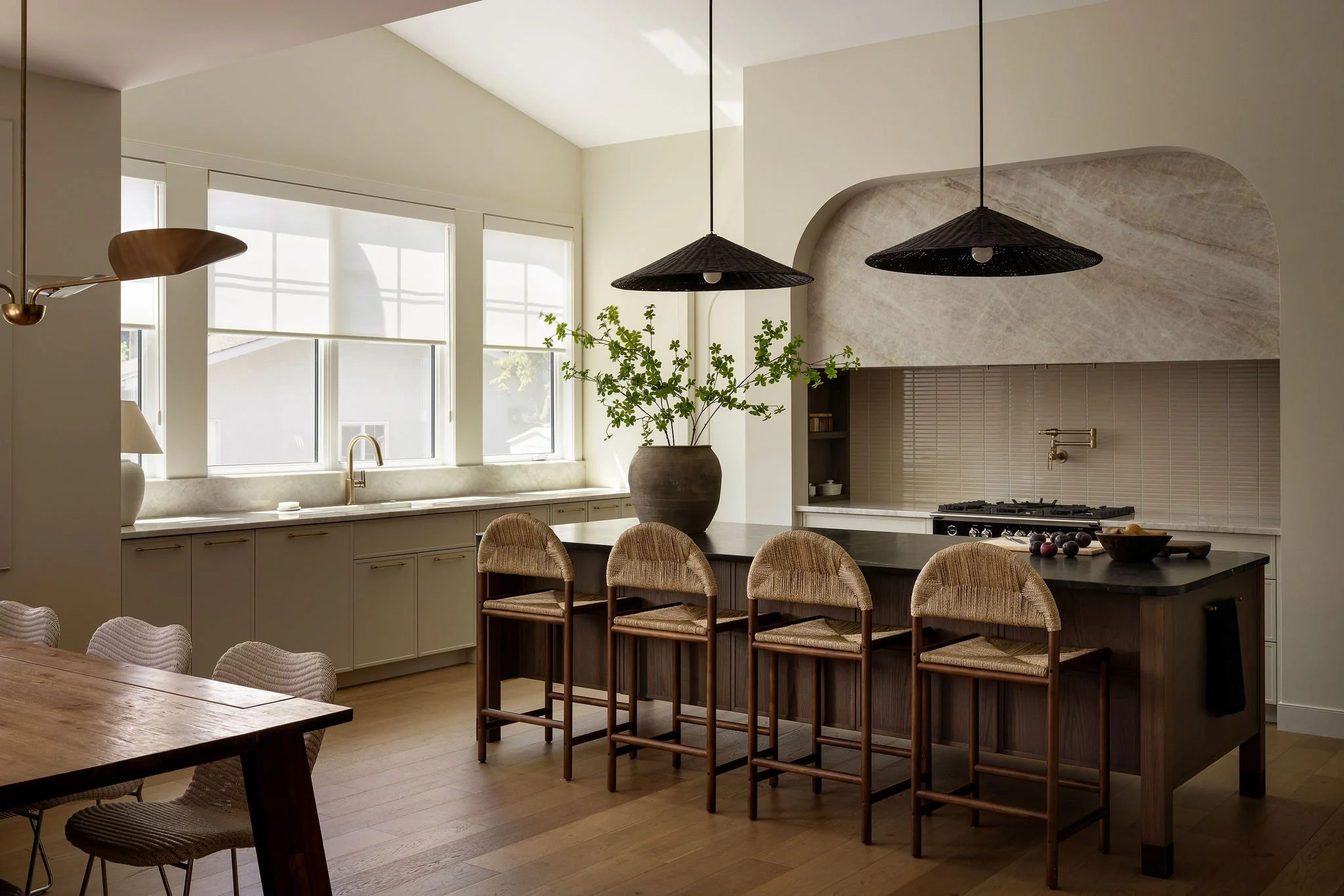 Modern kitchen with large windows, a kitchen island with bar stools, a gas stove, and a potted plant on the countertop. Black pendant lights hanging above the island and natural light filling the space.