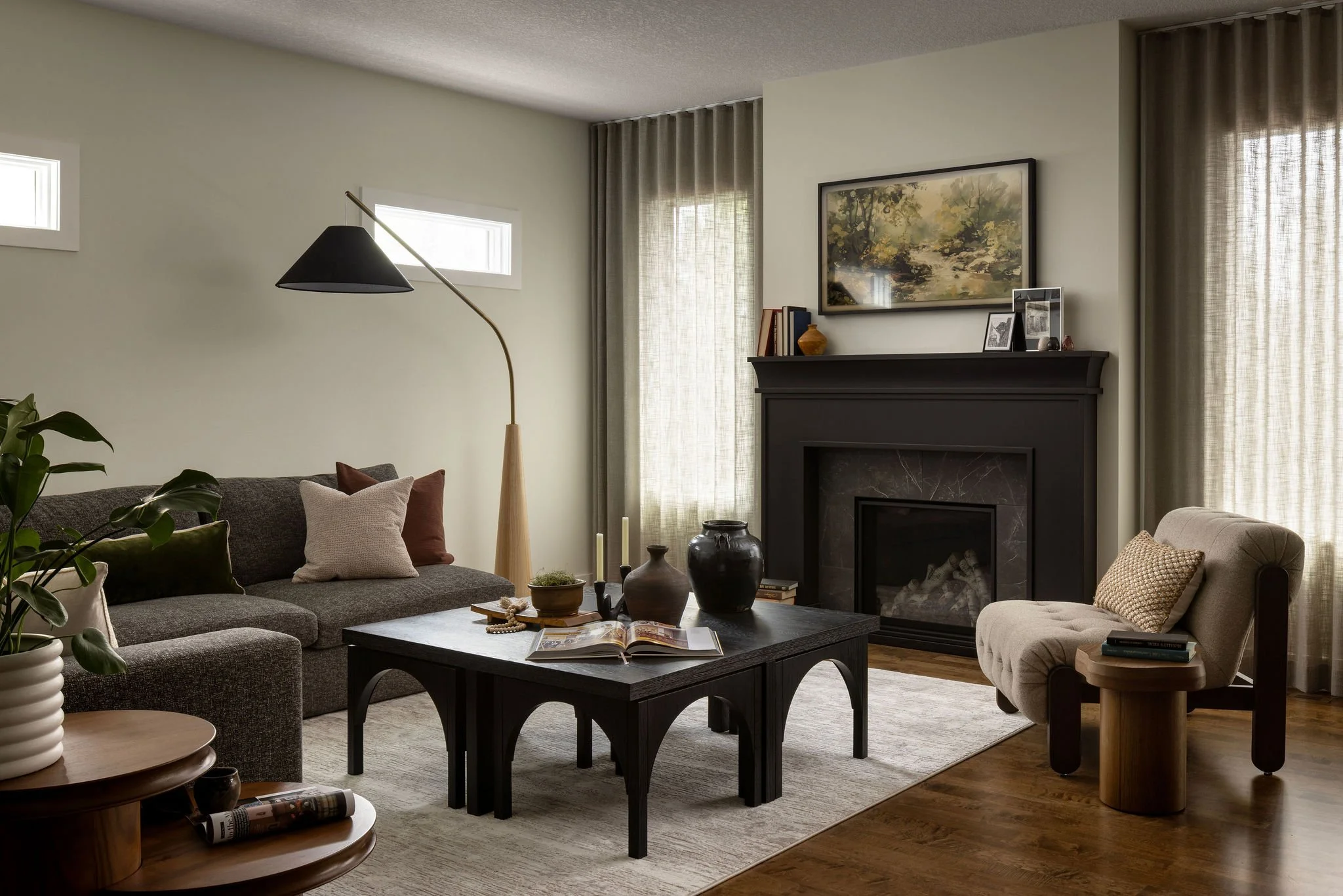 Living room with a gray sectional sofa, beige armchair, black coffee table, and fireplace. Decor includes plants, books, and framed photos, with natural light from windows and curtains.