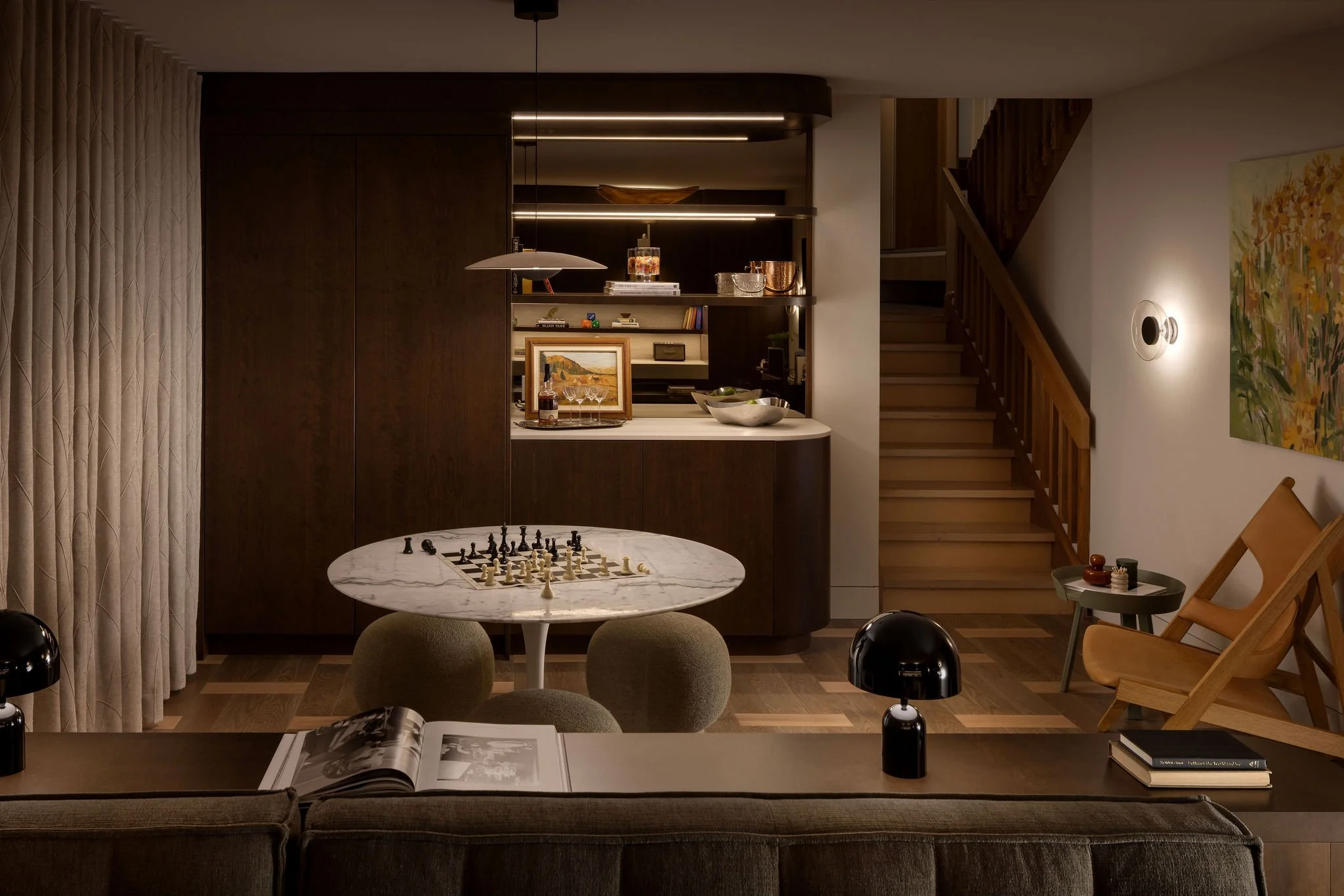 Interior of a modern living space with a marble-topped round dining table set for a chess game, a brown sofa with an open magazine, a black coffee bottle, in front of a dark wood built-in wall unit with shelves decorated with books, artwork, and decorative objects. To the right is a staircase with wooden steps and railing, and a wall-mounted light fixture next to colorful floral artwork.