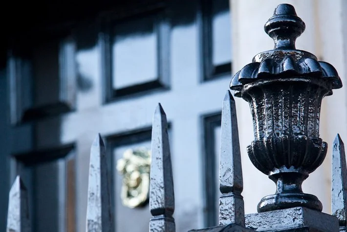 Close-up of a black wrought iron fence with spear-shaped tips in front of a building with windows and a blurred emblem on the wall.
