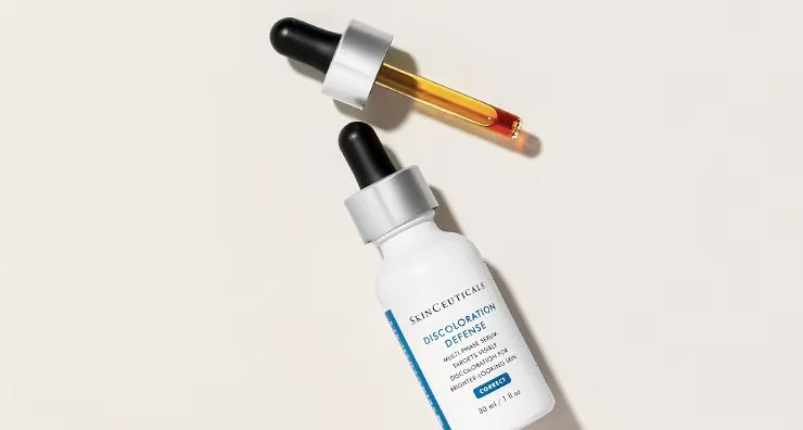 A skincare bottle labeled 'Discoloration Defense' with a dropper applicator laying nearby containing a yellow-orange liquid.