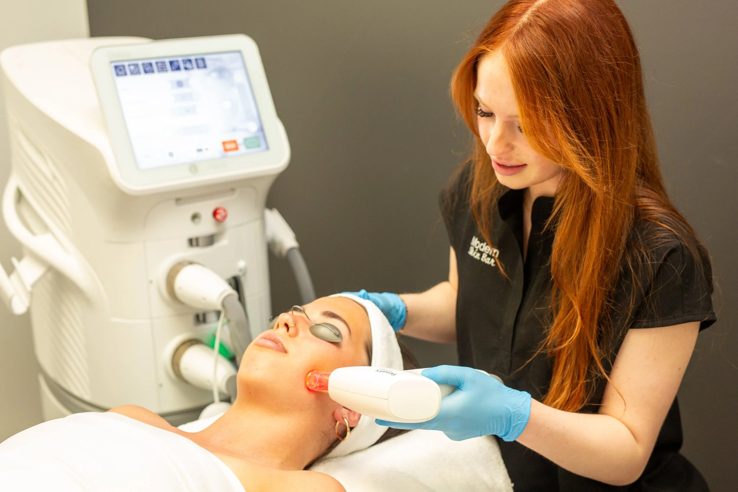 A woman receiving a facial treatment from a professional in a clinic with specialized equipment.
