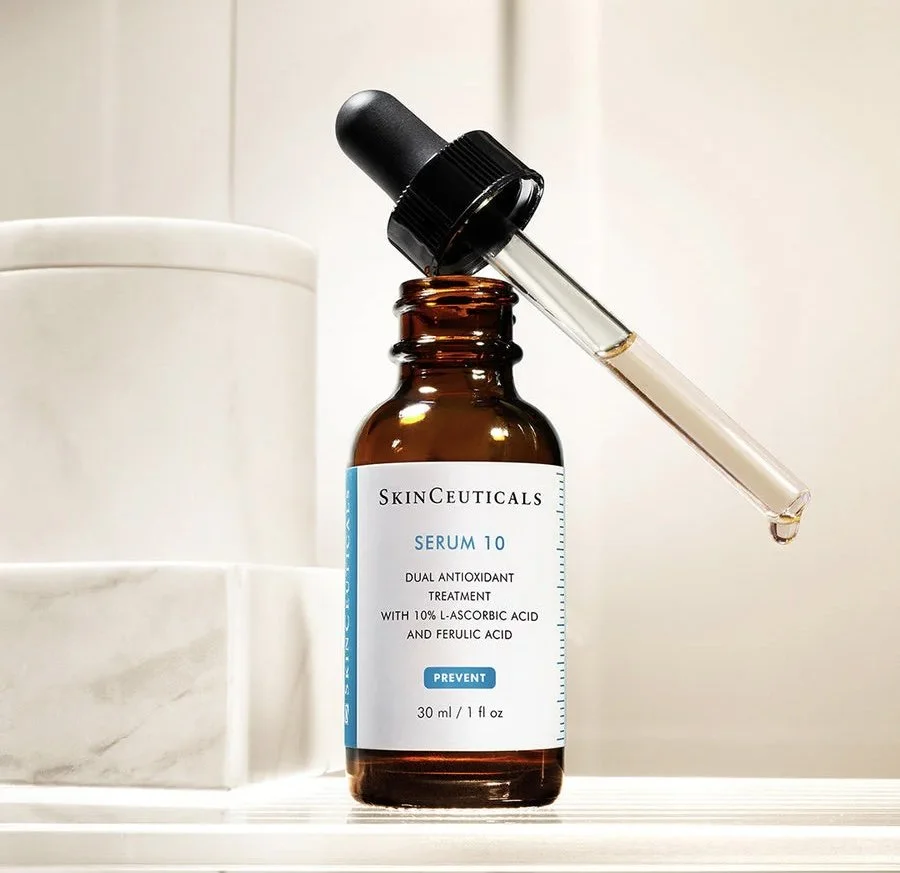 A brown glass dropper bottle labeled "SkinCeuticals Serum 10," with a black dropper cap, placed on a white surface, with a pipette containing a clear liquid, and white containers in the blurry background.
