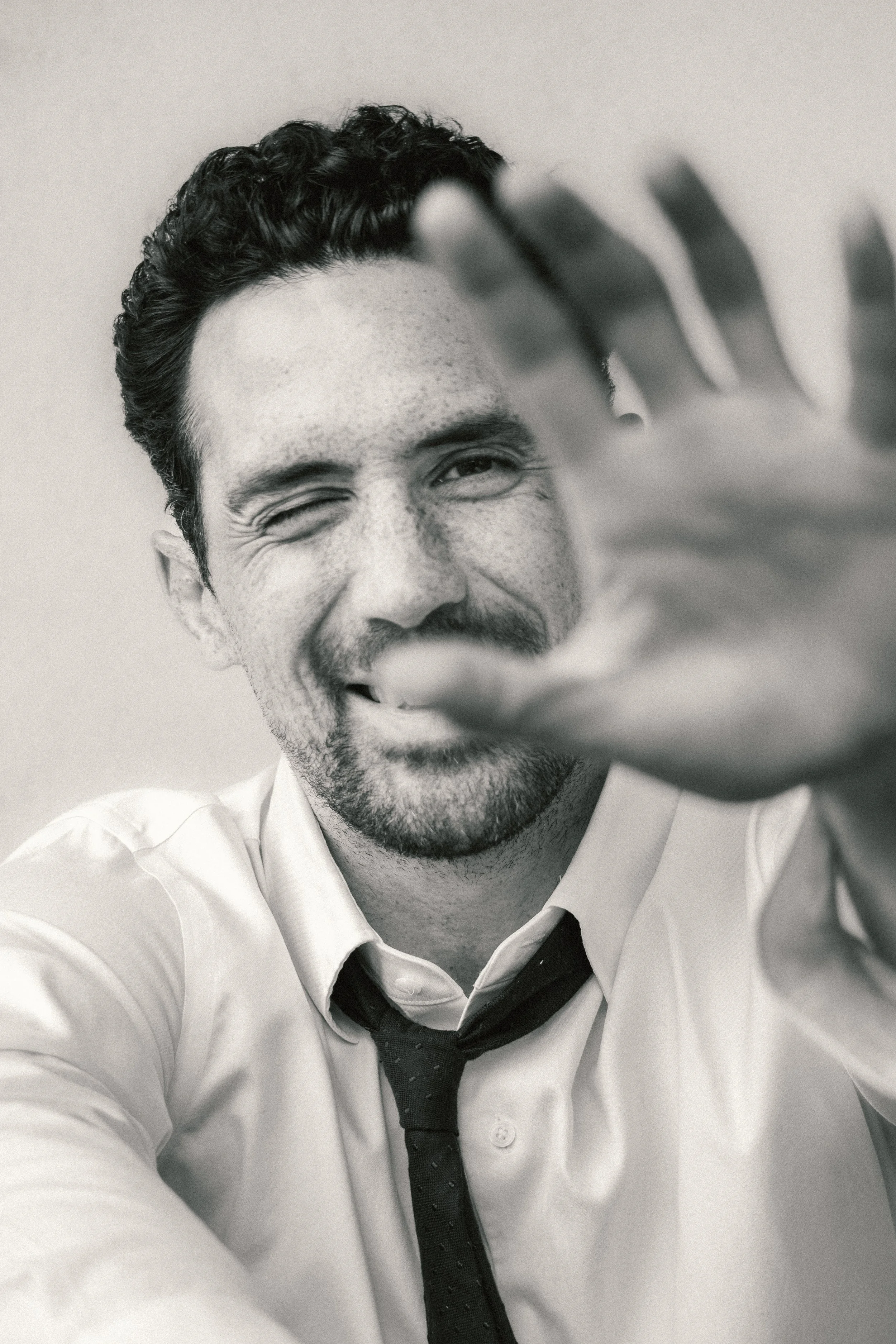 A black and white photo of a smiling man with curly hair, wearing a white shirt and a loosened tie, playfully covering part of his face with his hand.