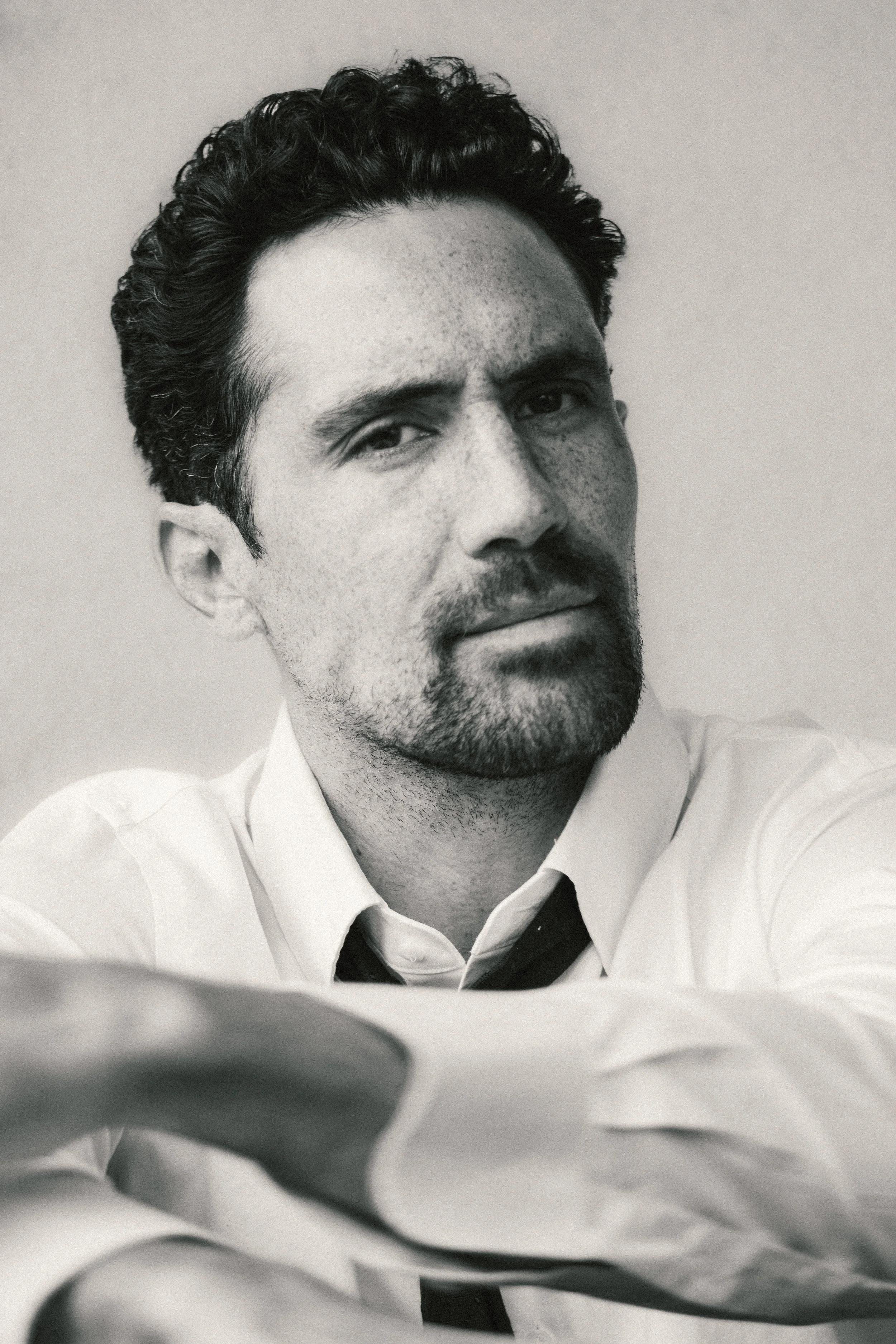 A black-and-white portrait of a man with curly dark hair, facial hair, and freckles, wearing a collared shirt, looking slightly to the side with a serious expression.