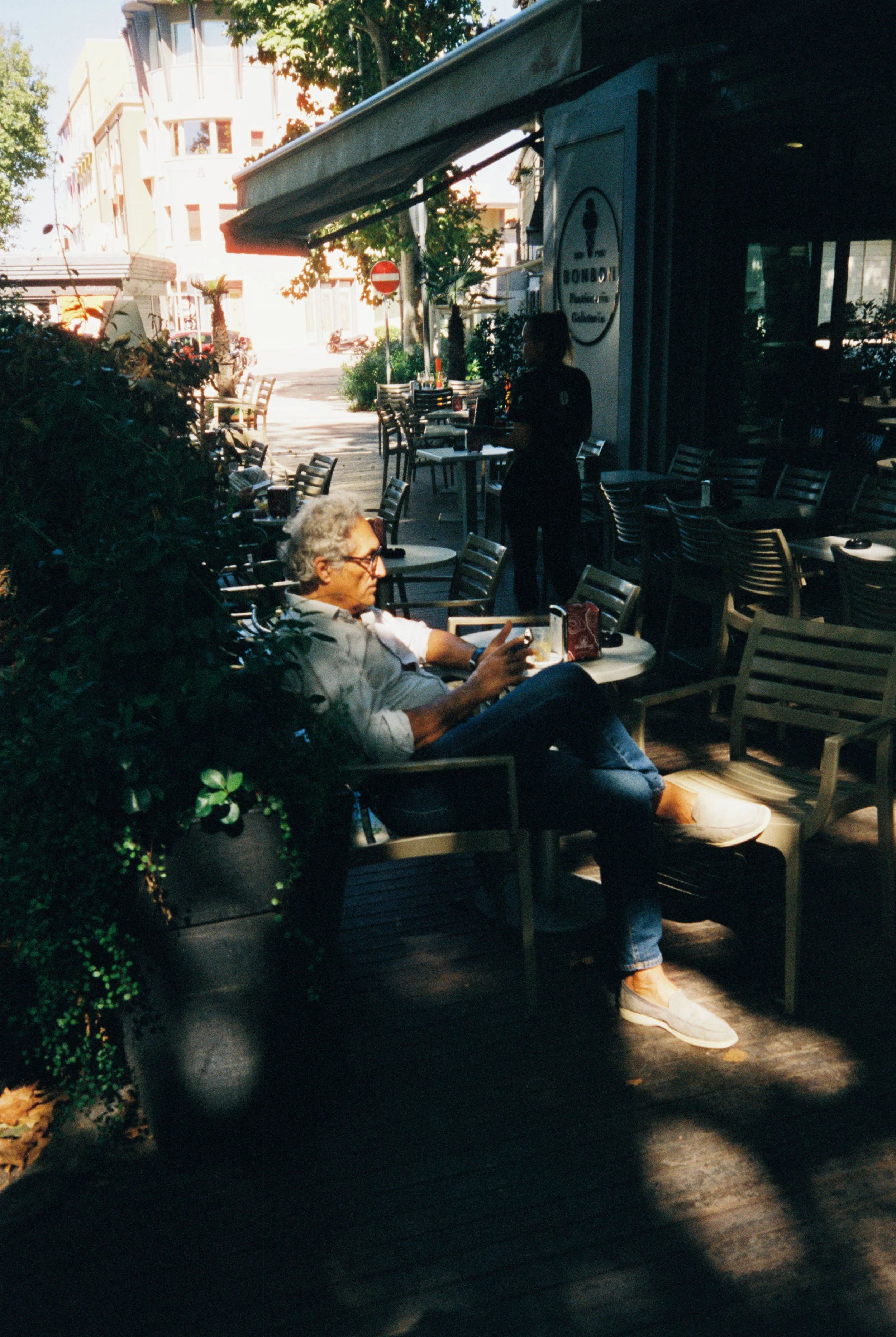 Ein Mann sitzt draußen auf einer Terrasse in der Sonne, liest auf seinem Smartphone, während eine Servicekraft im Schatten steht. Es gibt mehrere Tische und Stühle im Außenbereich eines Cafés.