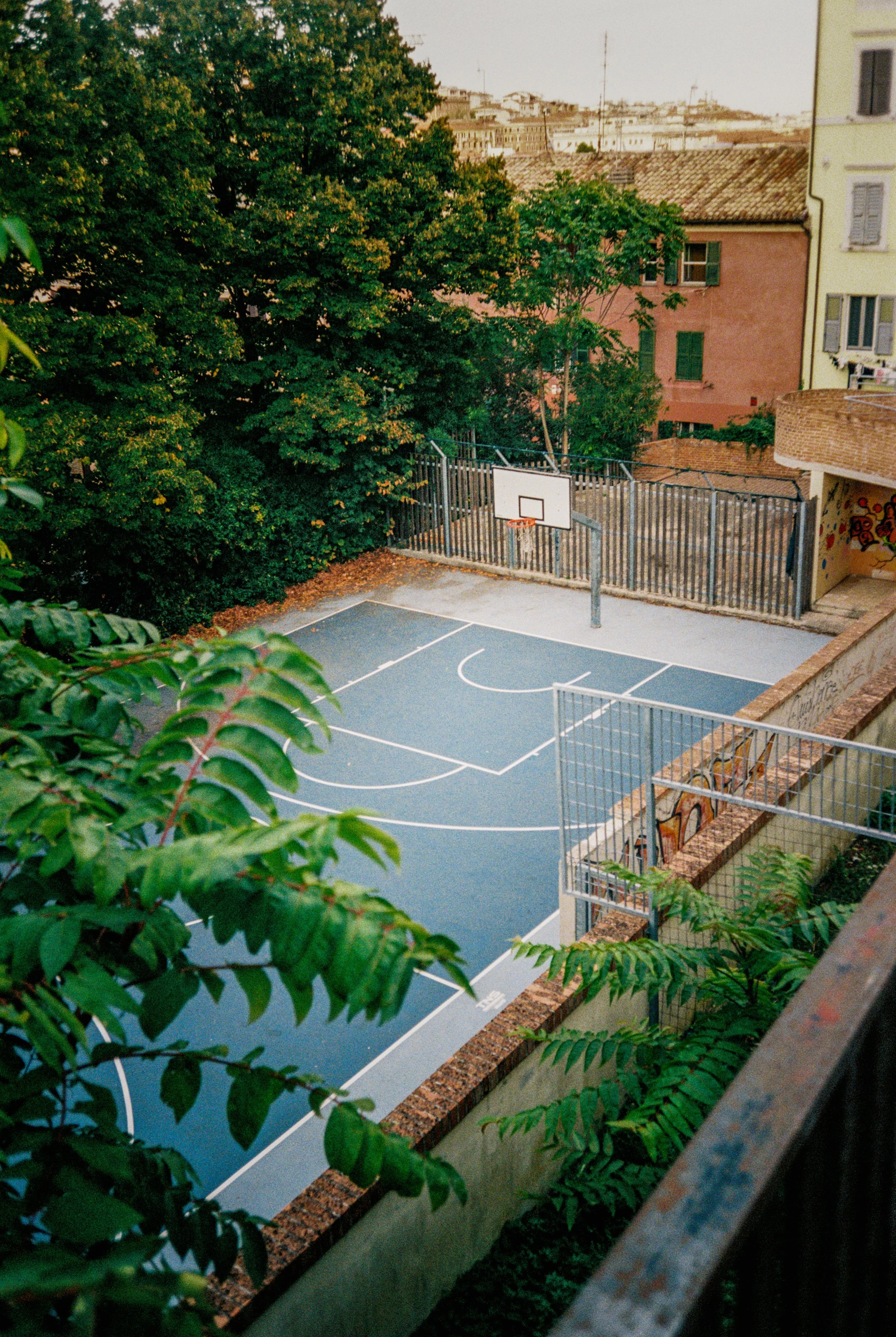 Ein leerer Basketballplatz in einer urbanen Gegend, umgeben von Bäumen und mehrstöckigen Gebäuden, aufgenommen bei Tageslicht.