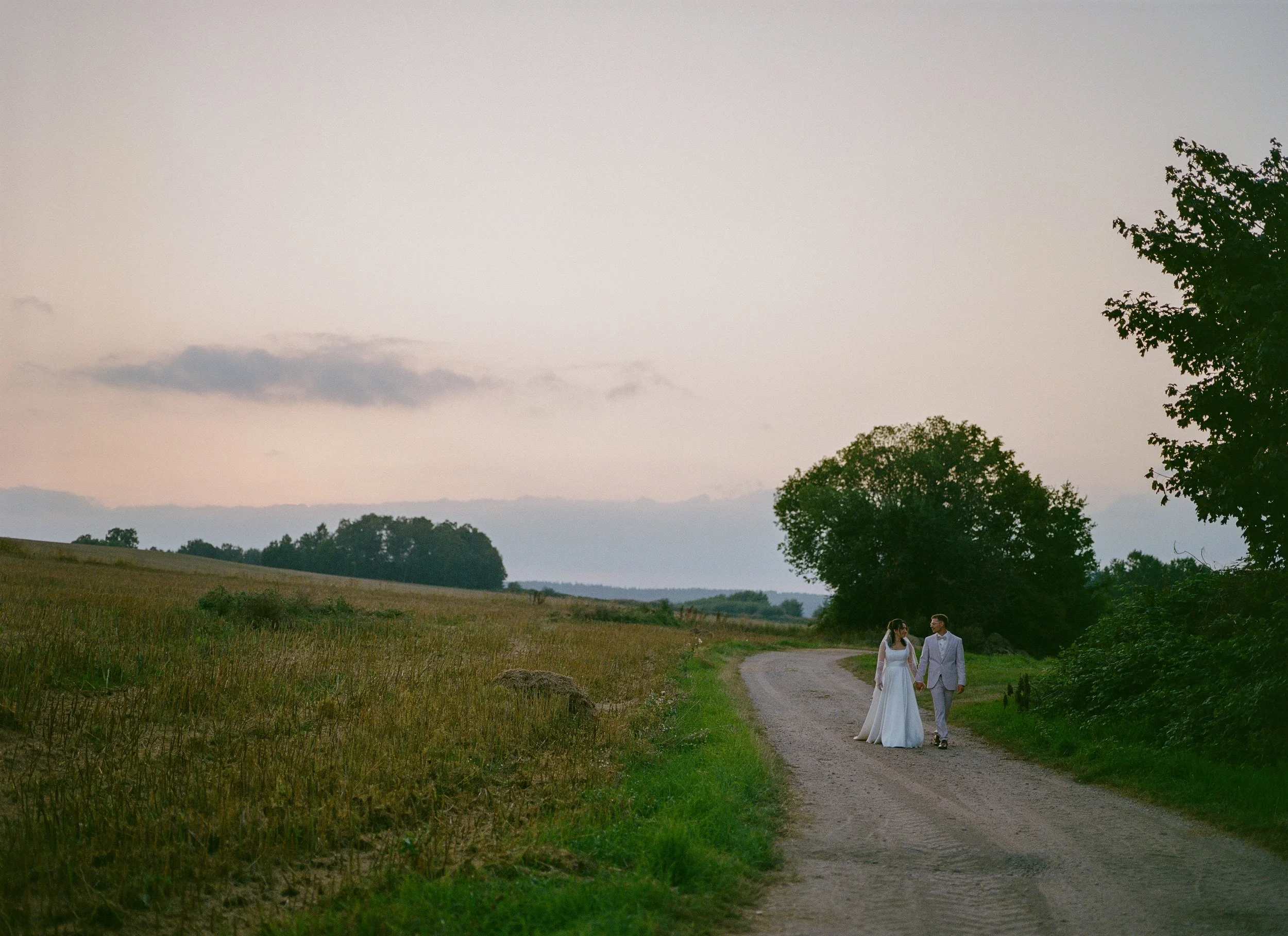 Ein frisch vermähltes Paar in Hochzeitskleidung spaziert Hand in Hand auf einem ländlichen Weg bei Sonnenuntergang, umgeben von offenen Feldern und Bäumen.