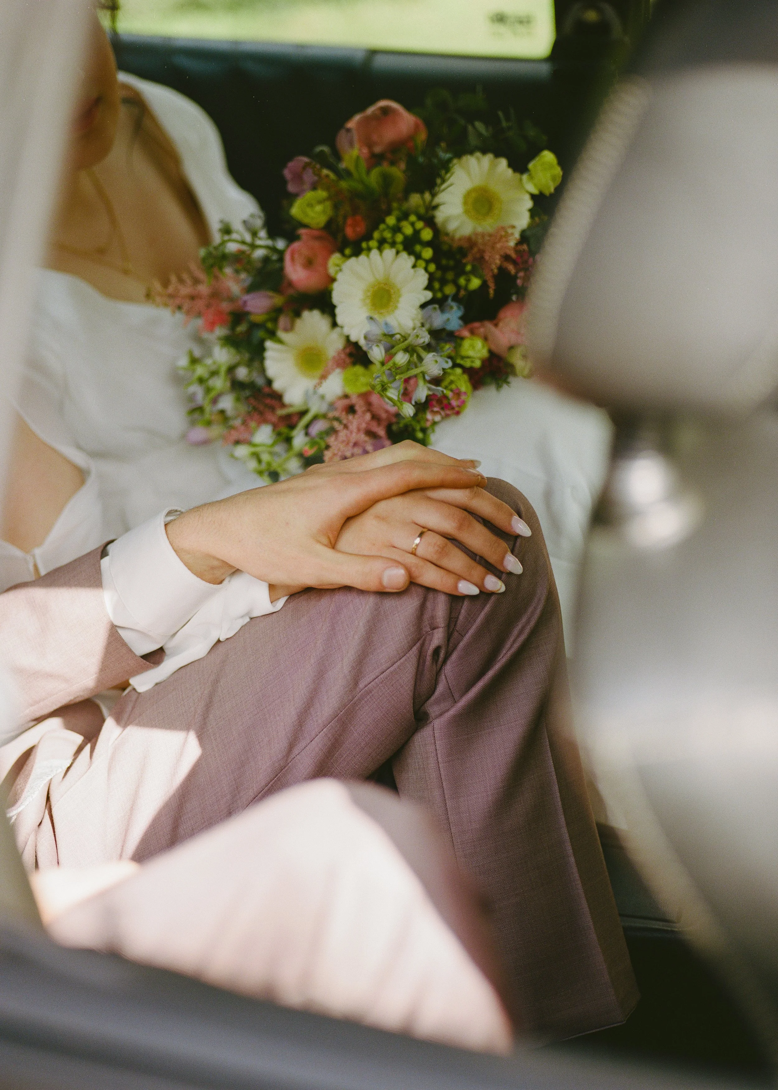 Nahaufnahme einer Hand mit Ehering, die auf einem Knie liegt, im Hintergrund ein Brautstrauß mit weißen, rosa und lila Blumen, Szene im Auto, vermutlich Hochzeitsfeier.