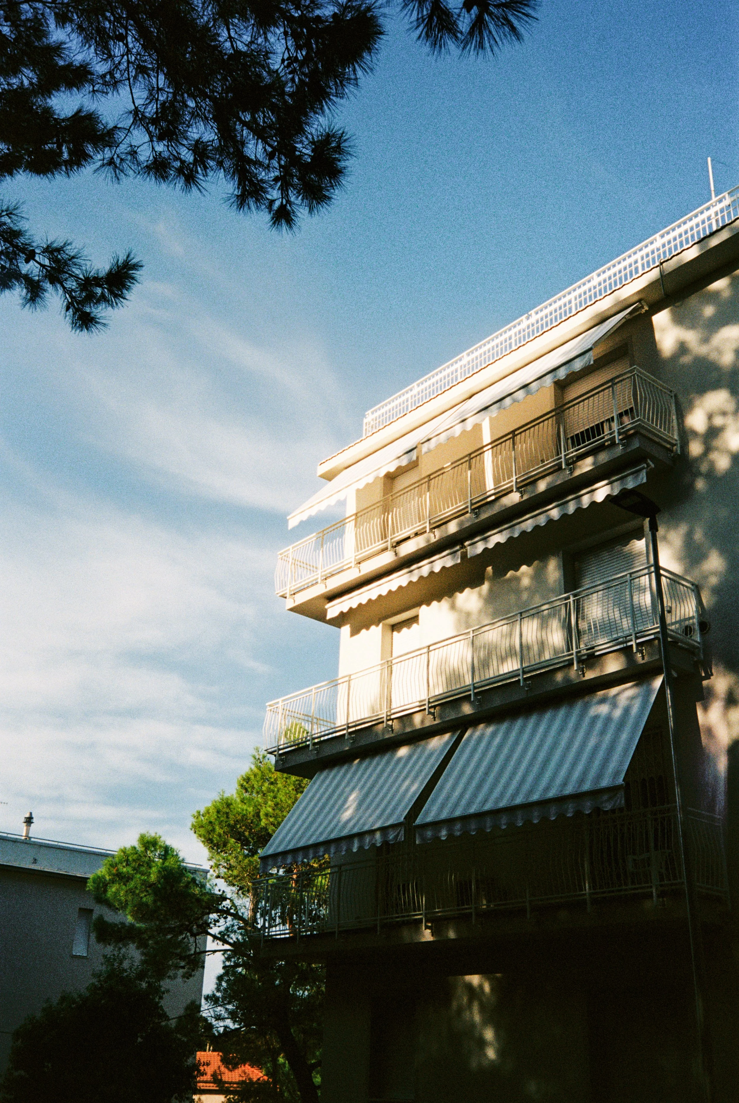 Mehrstöckiges Apartmentgebäude mit Balkonen und Markisen, umgeben von Bäumen, bei Sonnenuntergang.