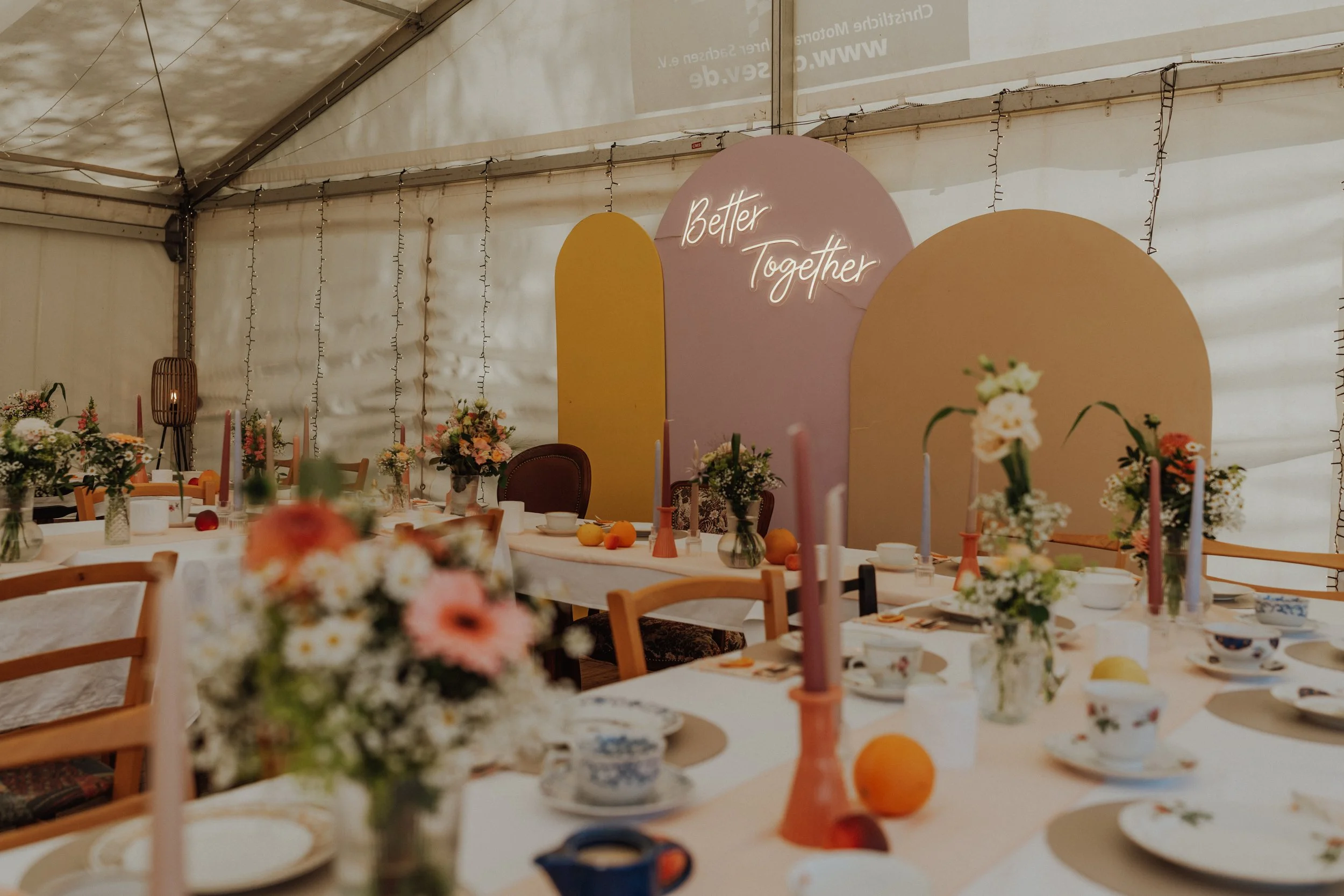 Ein festlich gedeckter Tisch mit Blumen, Kerzen und Geschirr in einem Zelt, im Hintergrund eine Wand mit einem Neon-Schild mit der Aufschrift 'Better Together'.