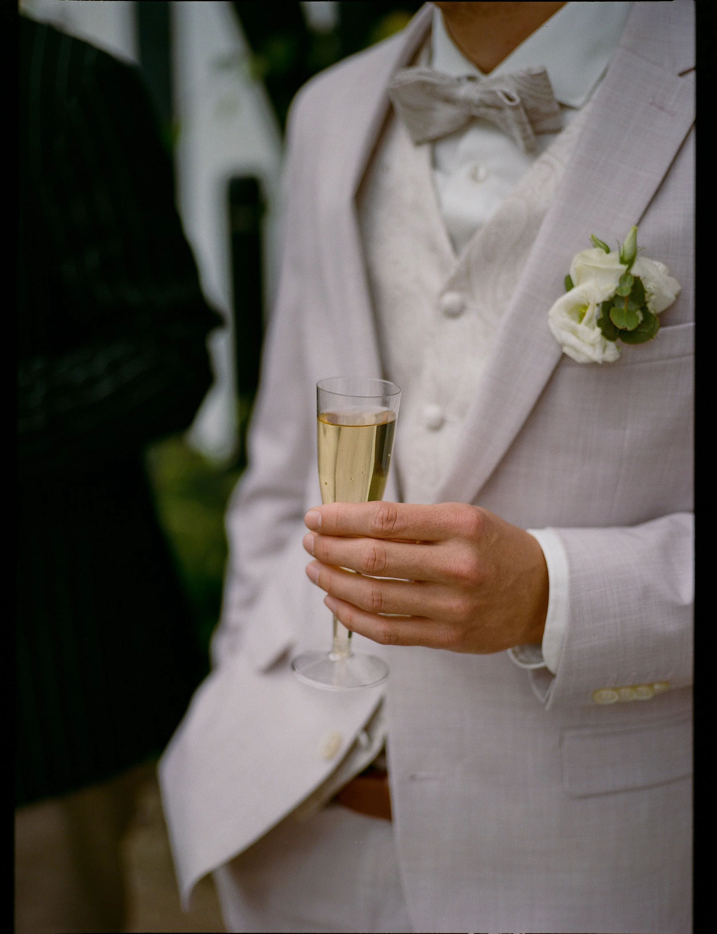 Mit einem schwarzen Anzug und einer weißen Fliege hält eine Person ein Glas Champagner bei einer Hochzeit.