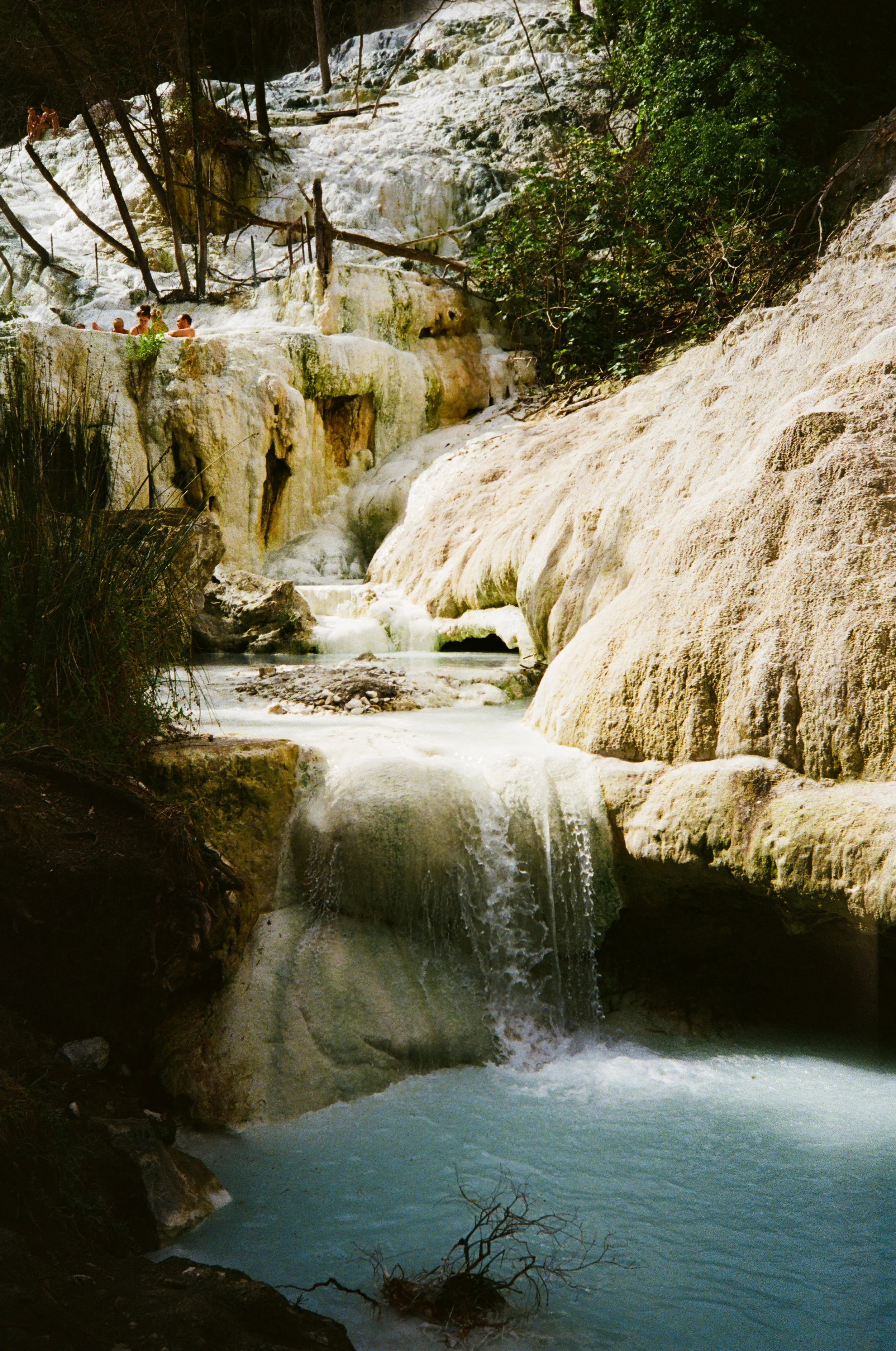 Ein Wasserfall in einer natürlichen Umgebung mit Felsen, Wasser und Bäumen, einige Personen sitzen im Wasser im oberen Bereich des Wasserfalls.