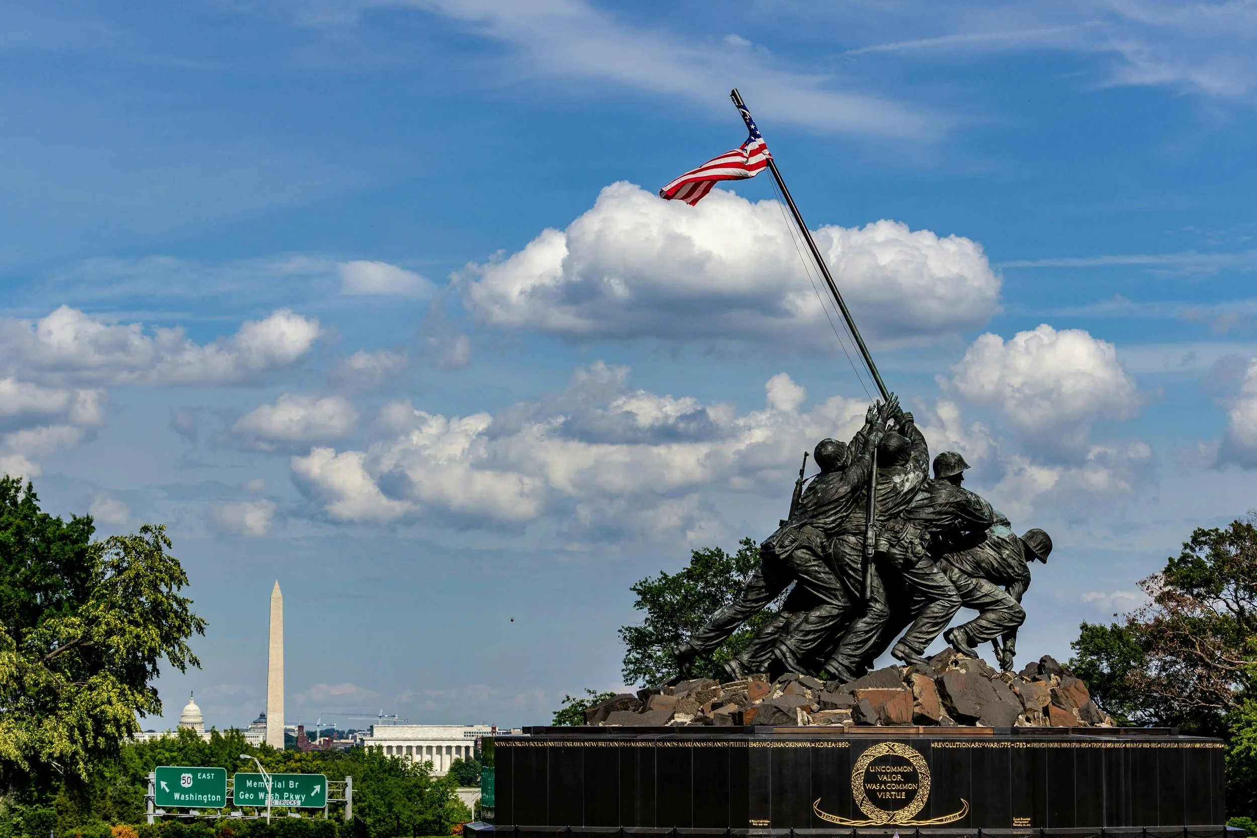Soldiers during World War II lift the American flag in Iwo Jima. The photo is of the statue in Washington, and shows theWashington monument, capitol, and Lincoln Memorial, on a sunny day with blue skies and white puffy clouds.