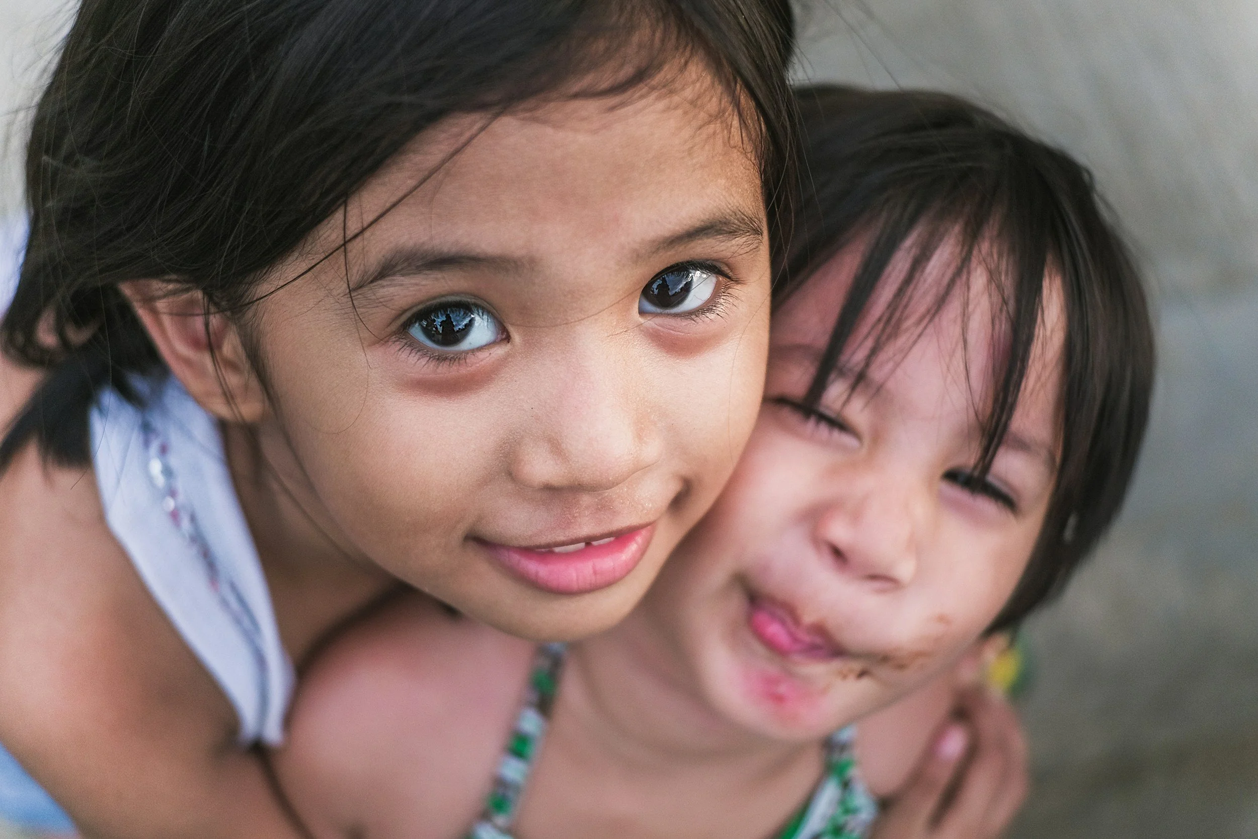 Two young girls with dark hair smiling and embracing each other outdoors.