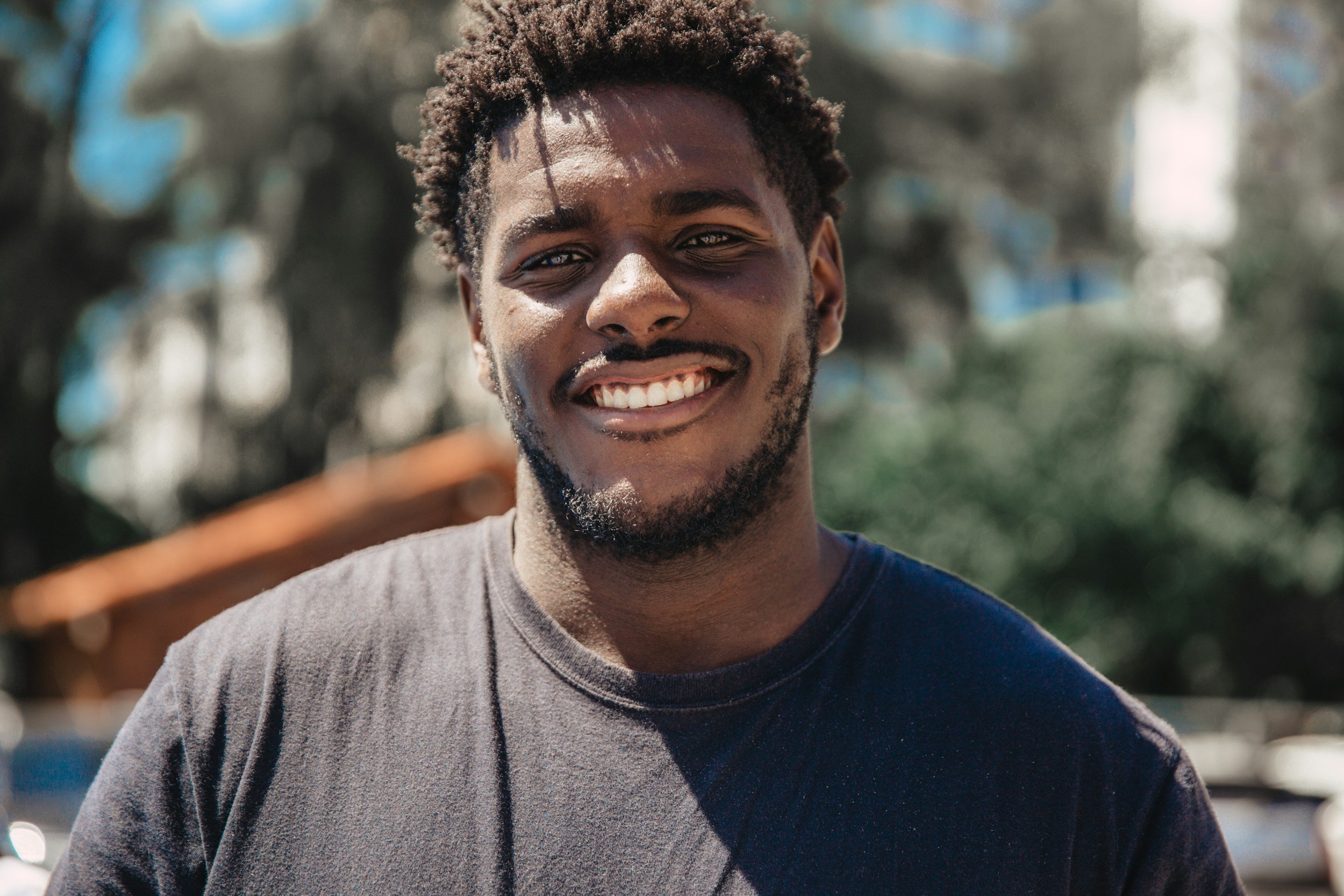 A young man with short curly hair and a beard smiling outside on a sunny day, wearing a dark t-shirt, with blurred trees and buildings in the background.