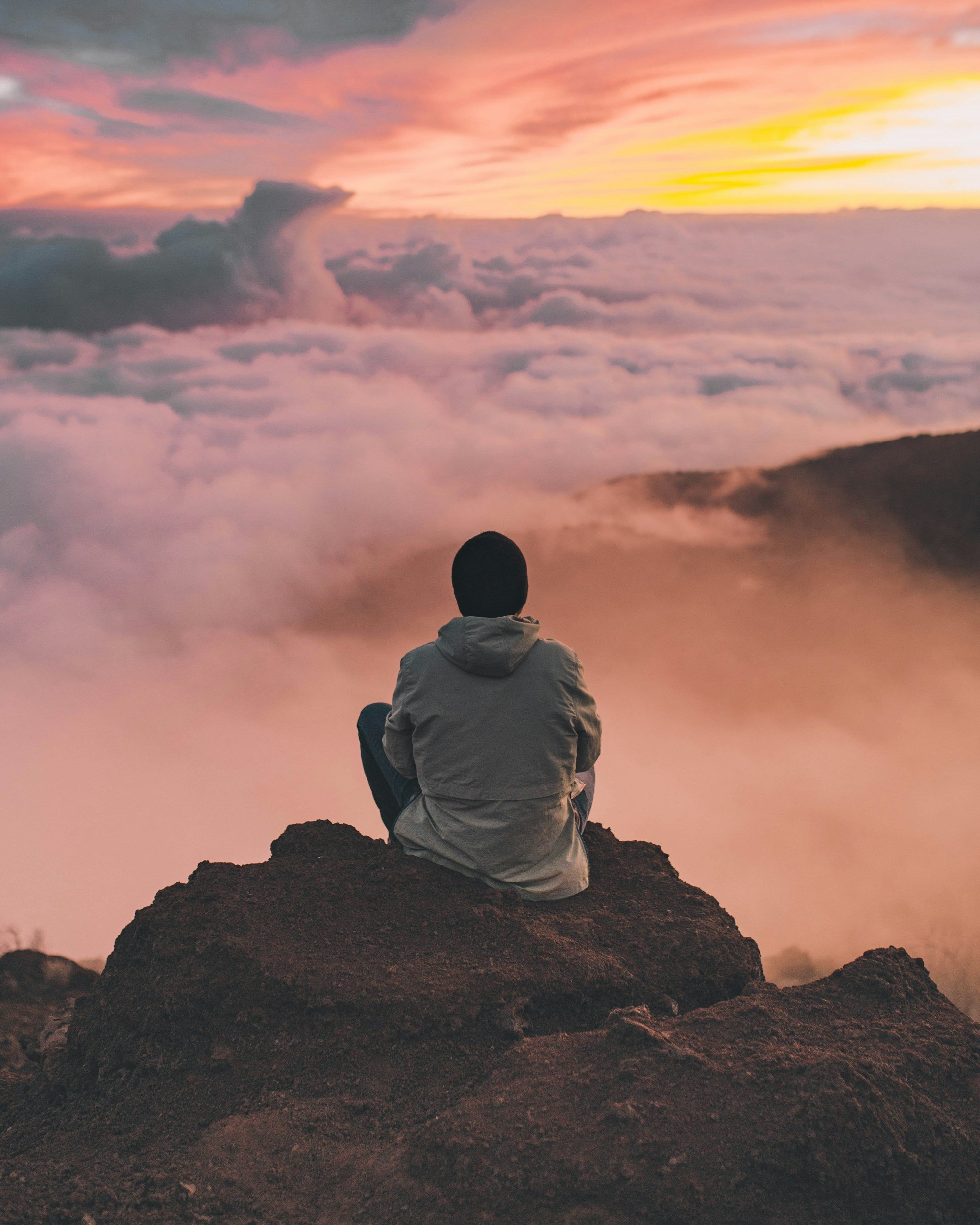 A person sitting on a rock, facing a sky filled with pink and orange clouds during sunset or sunrise, overlooking a sea of clouds below a mountain.