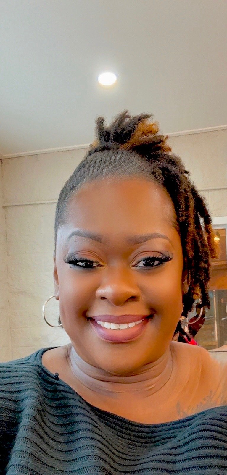 A woman smiling with makeup and hoop earrings, wearing a black top with textured fabric, indoors with a ceiling light and beige wall in the background.
