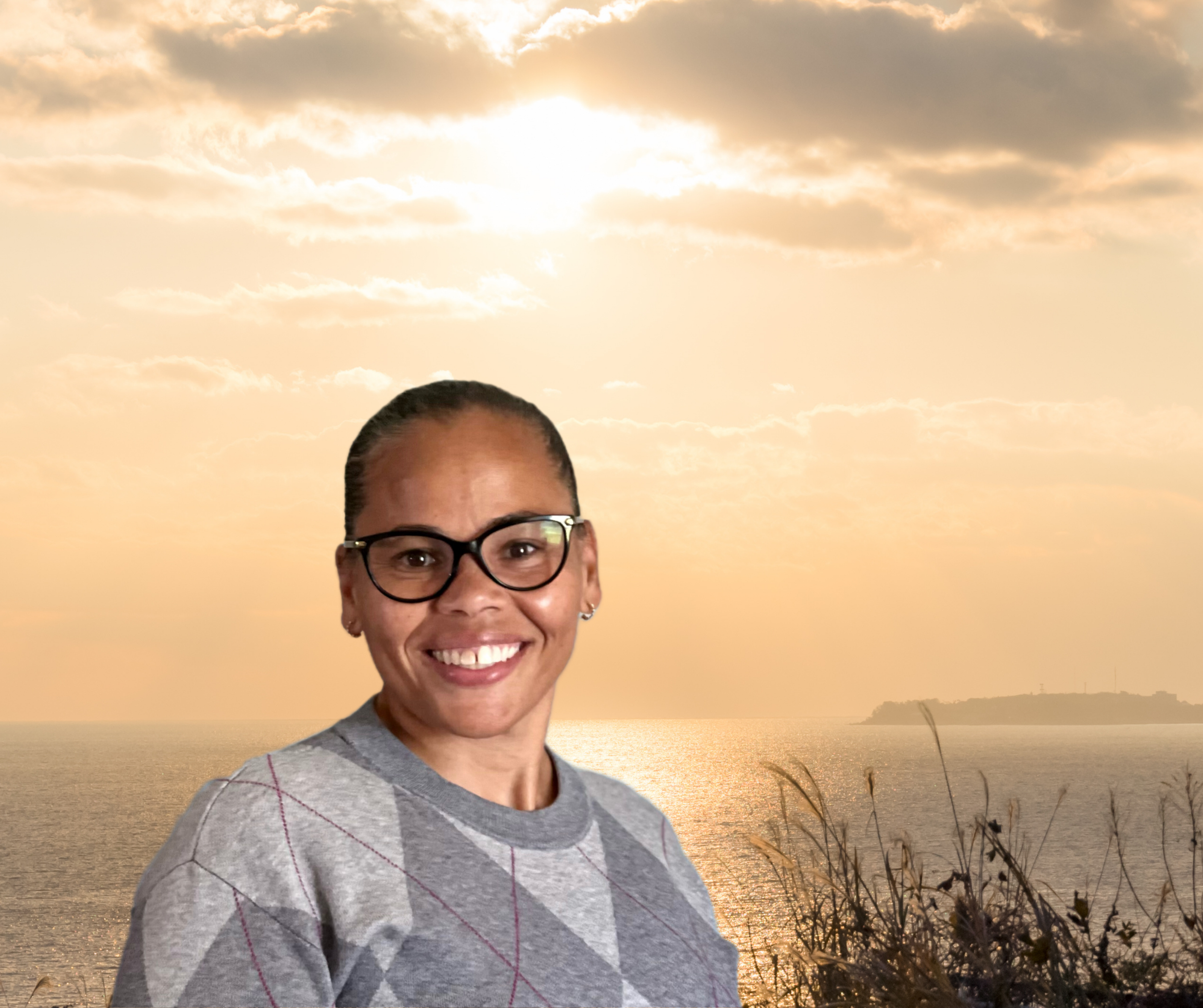 A smiling woman with glasses and earrings standing outdoors at sunset near the ocean with clouds and a distant landmass.