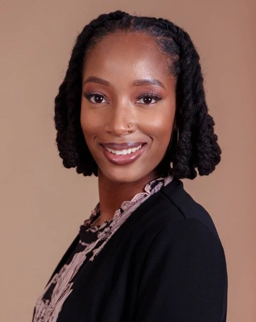 A woman with shoulder-length curly black hair and a nose piercing, smiling, wearing a black blazer and a patterned blouse, against a beige background.