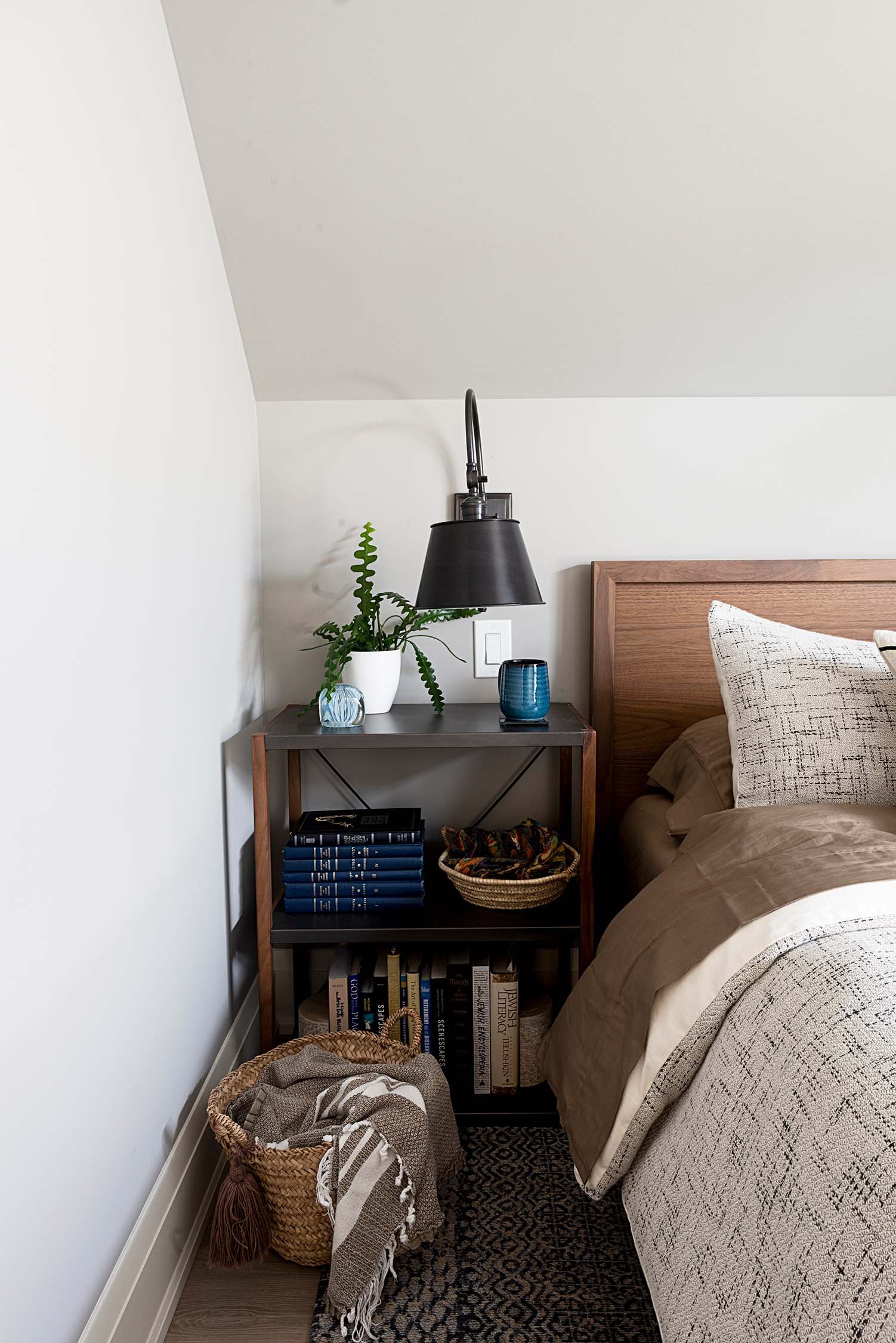 Bedroom corner with a wooden headboard, nightstand, black wall lamp, potted plant, blue mug, books, basket with textiles, and patterned bedding.