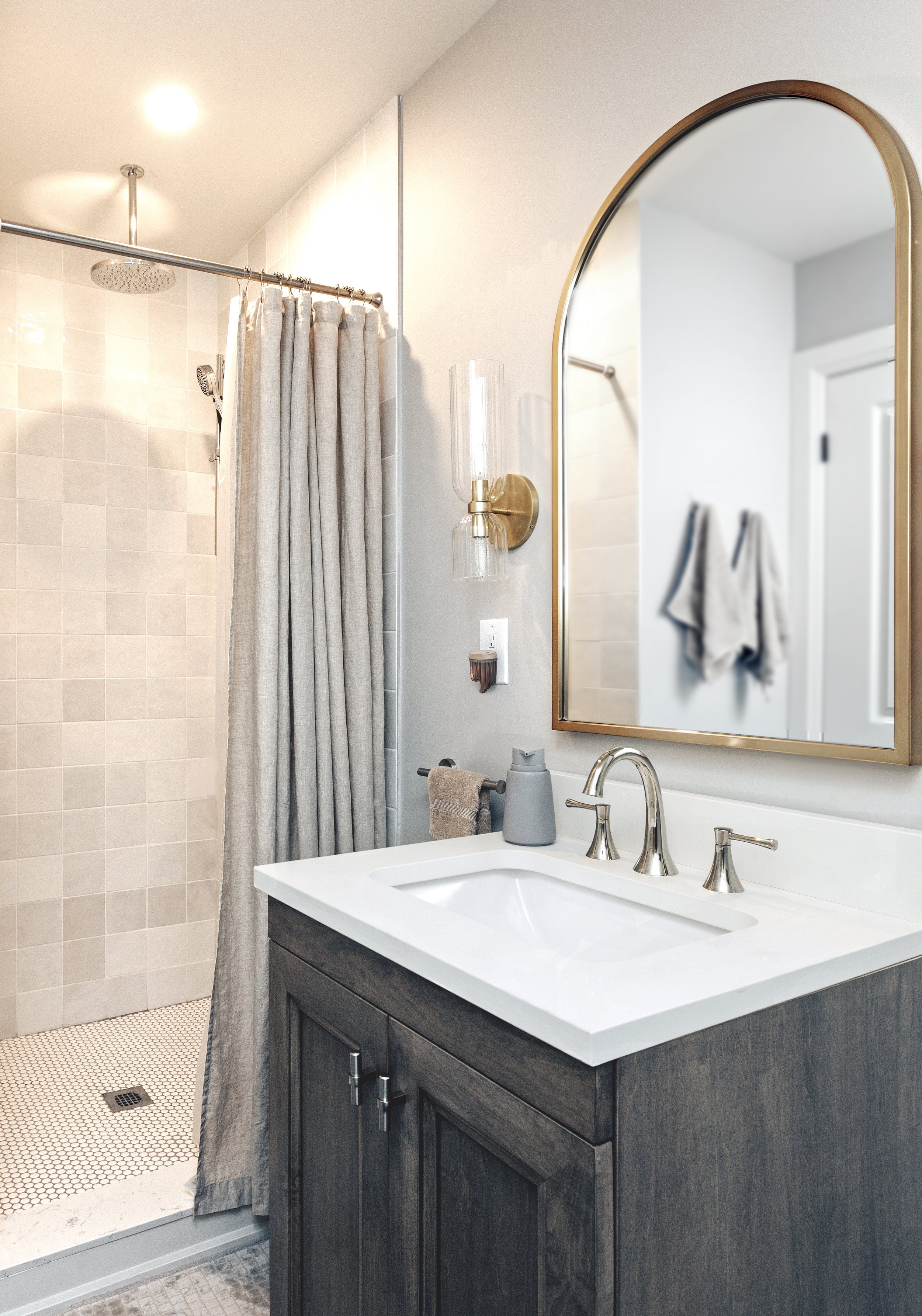 Modern bathroom with gray vanity sink and arched mirror, featuring a glass shower with beige curtain and wall-mounted light fixture.