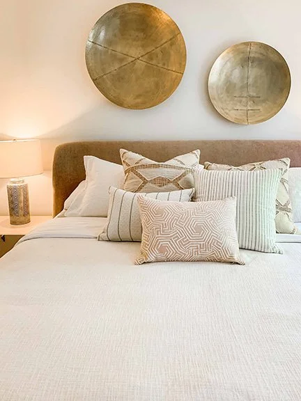 A neatly made bed with multiple decorative pillows, a beige headboard, with two round metallic wall art pieces above it, and a lamp on a nightstand on the left side.