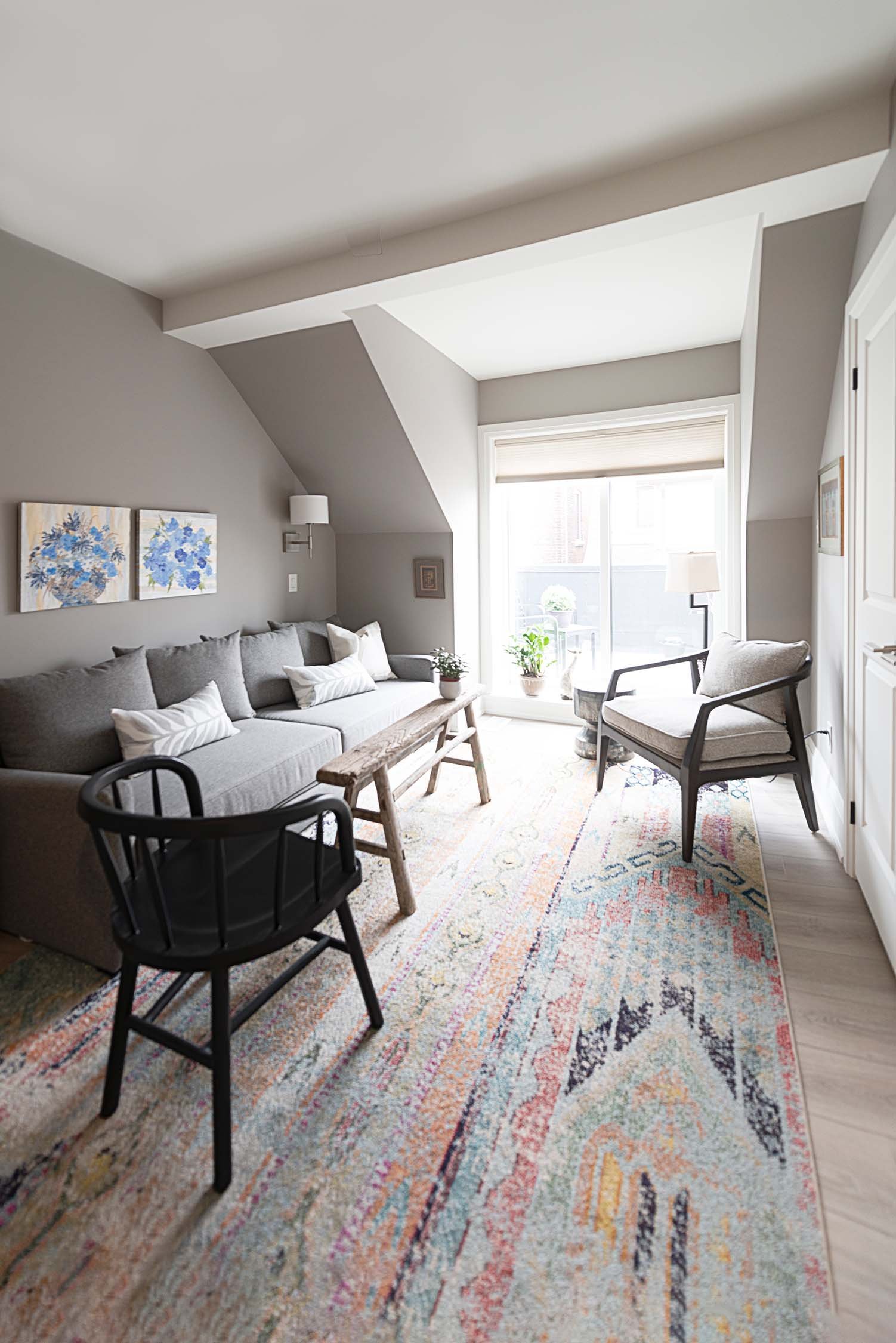 Modern living room with gray sofa, patterned rug, two chairs, a wooden coffee table, and wall art. Sunlight streams through a large window.