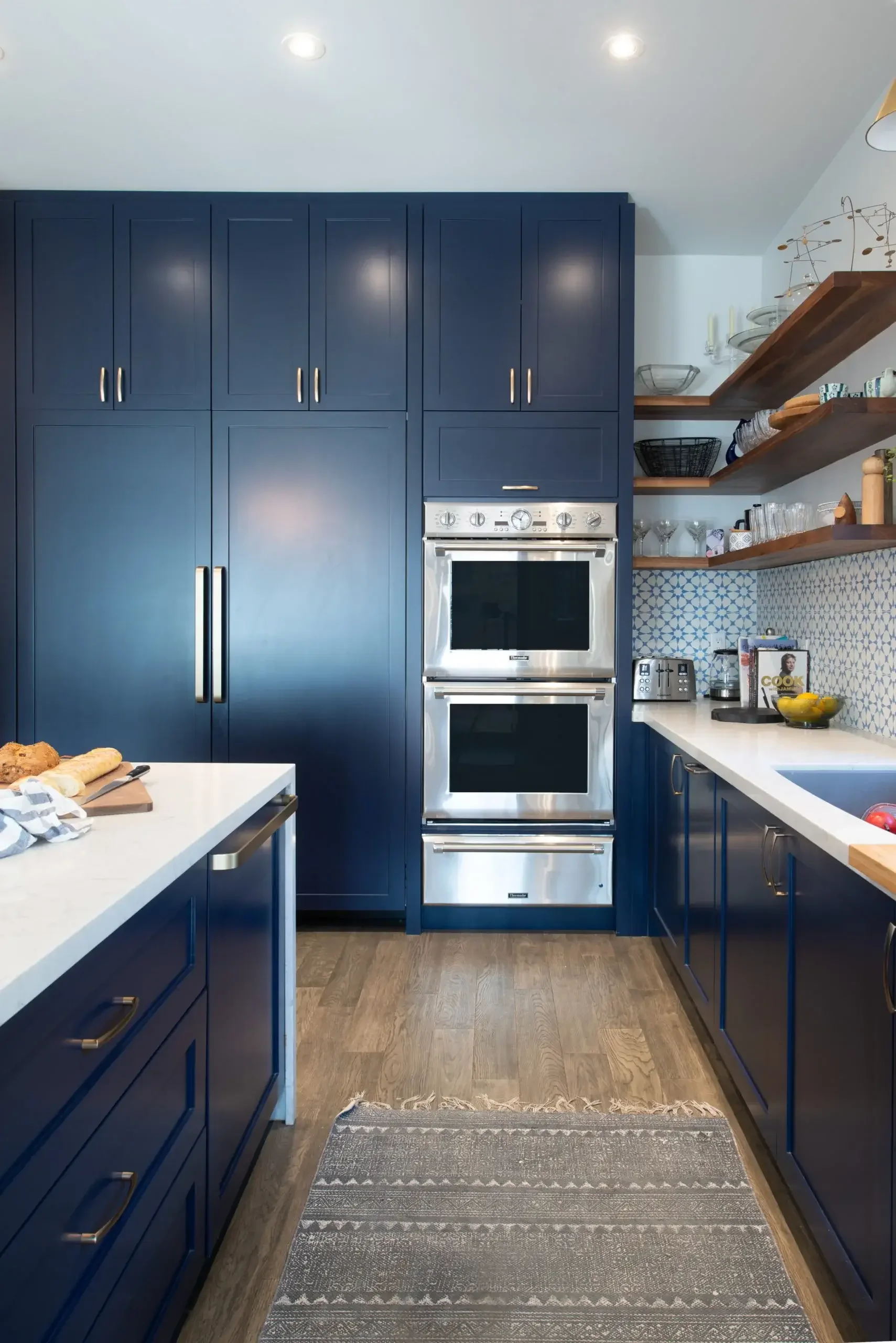 Modern kitchen with blue cabinets, stainless steel oven, open wooden shelves, and a white countertop. Contains kitchen items and bread on a cutting board.