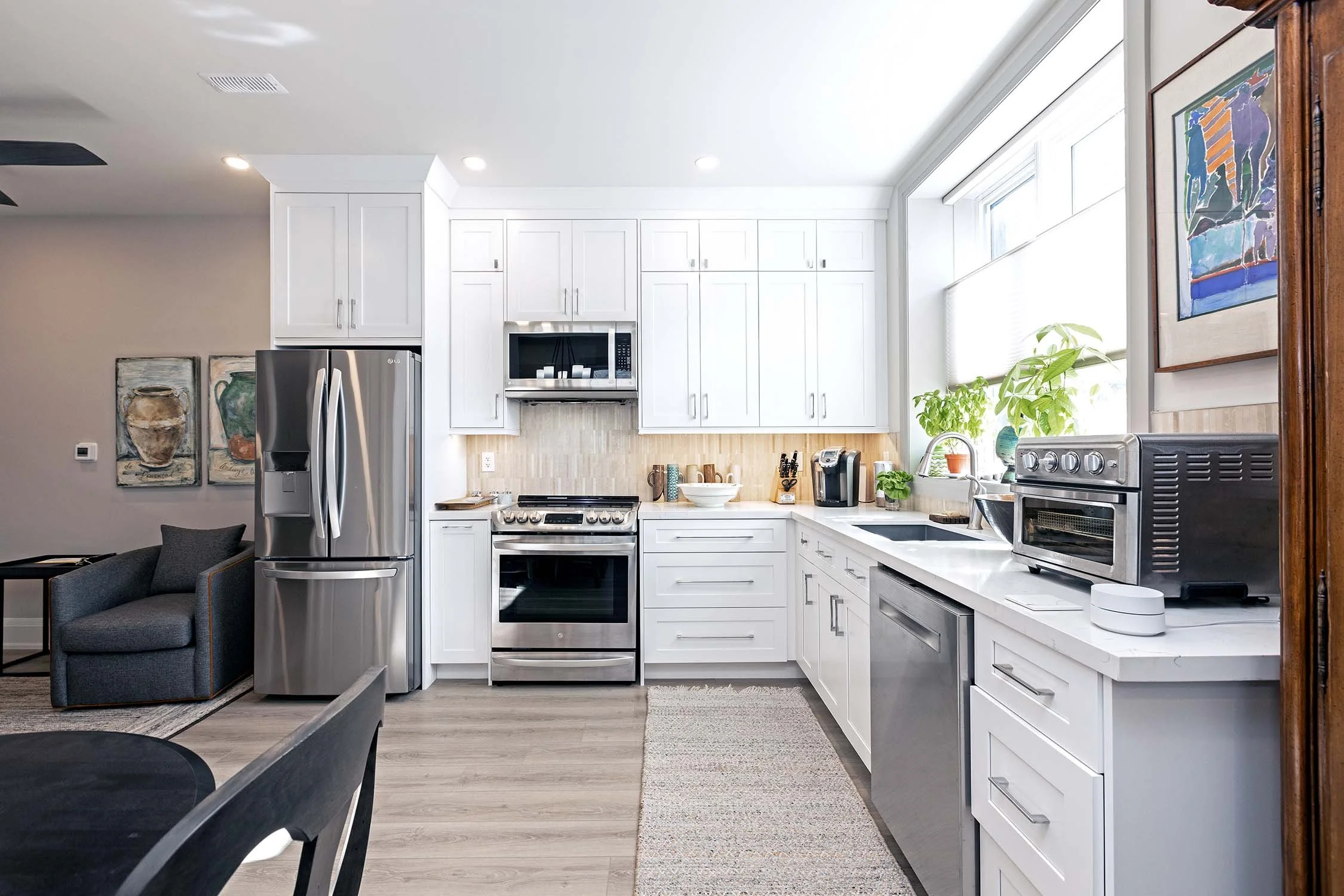Modern kitchen with stainless steel appliances, white cabinetry, and a window with plants. Features a refrigerator, oven, microwave, and countertop with a sink and toaster oven.