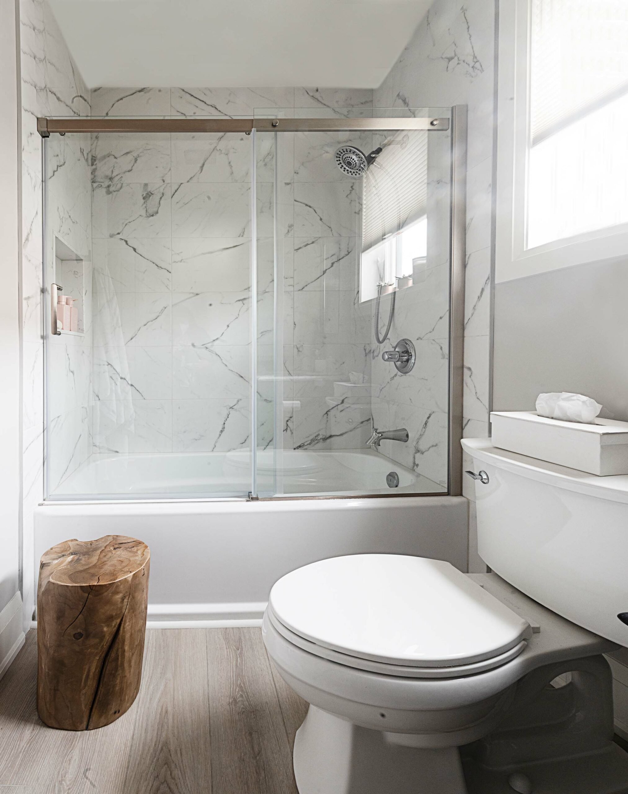 Modern bathroom with a white toilet, wooden stool, and bathtub with sliding glass doors. Marble walls and light wood floor.