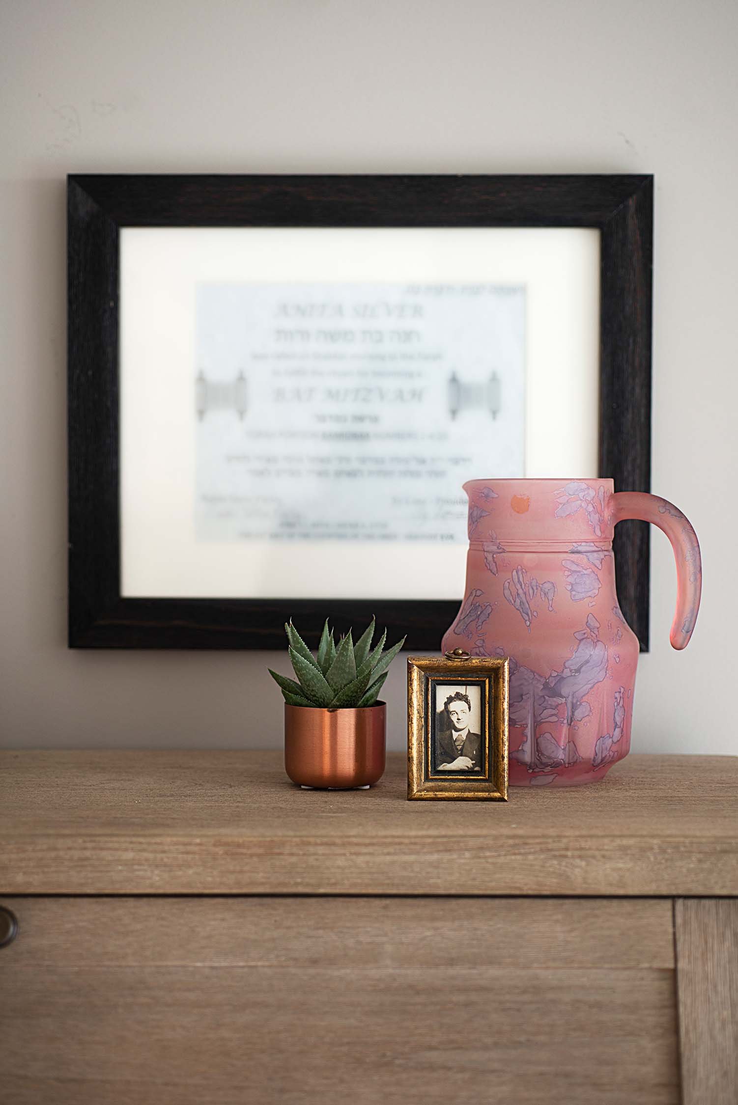 Wooden surface with a small green potted plant, a framed photograph, a pink decorative pitcher, and a blurred framed document on the wall.