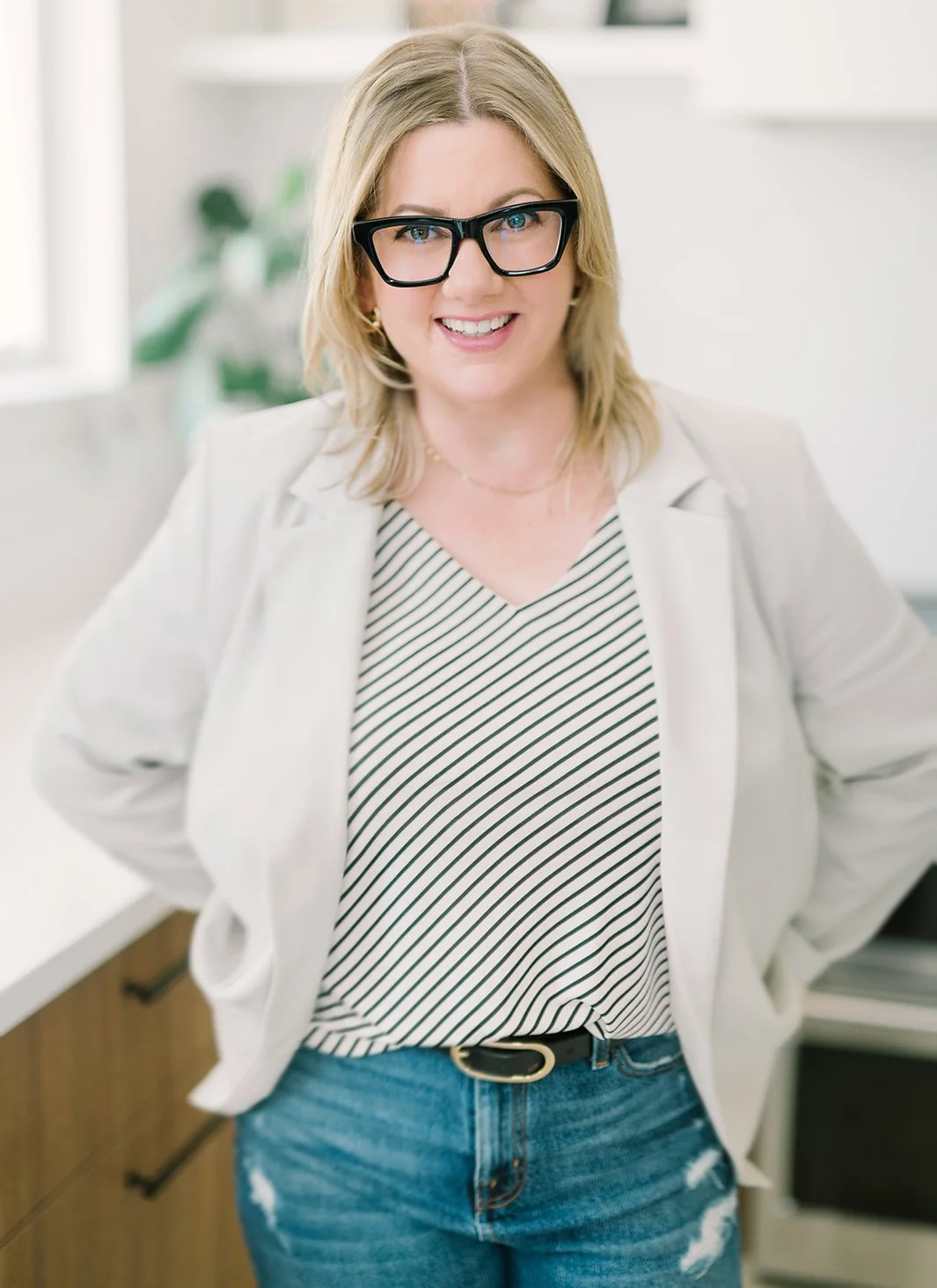 Person wearing glasses, striped top, light blazer, and jeans, smiling in a kitchen setting.