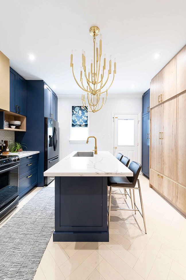 Modern kitchen with navy blue and light wood cabinetry, white marble island with gold faucet, black bar stools, and a gold chandelier.