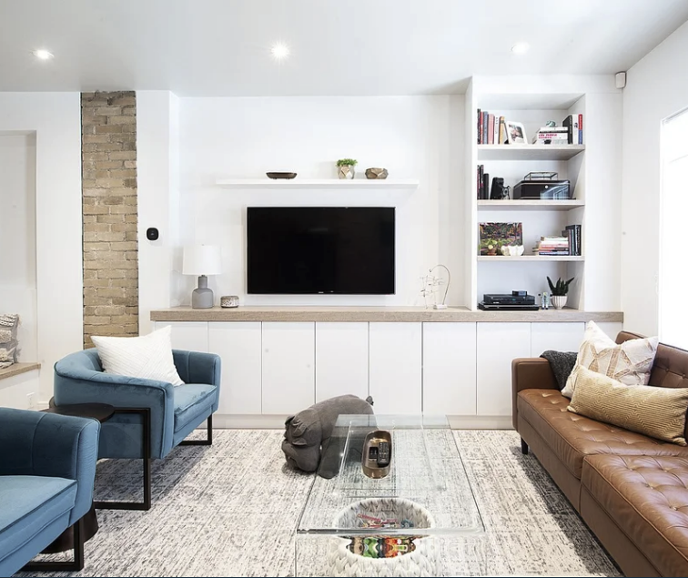 Modern living room with blue armchairs, a brown leather couch, a glass coffee table, built-in shelves with books and decor, a wall-mounted TV, and wall art with a colorful owl illustration.