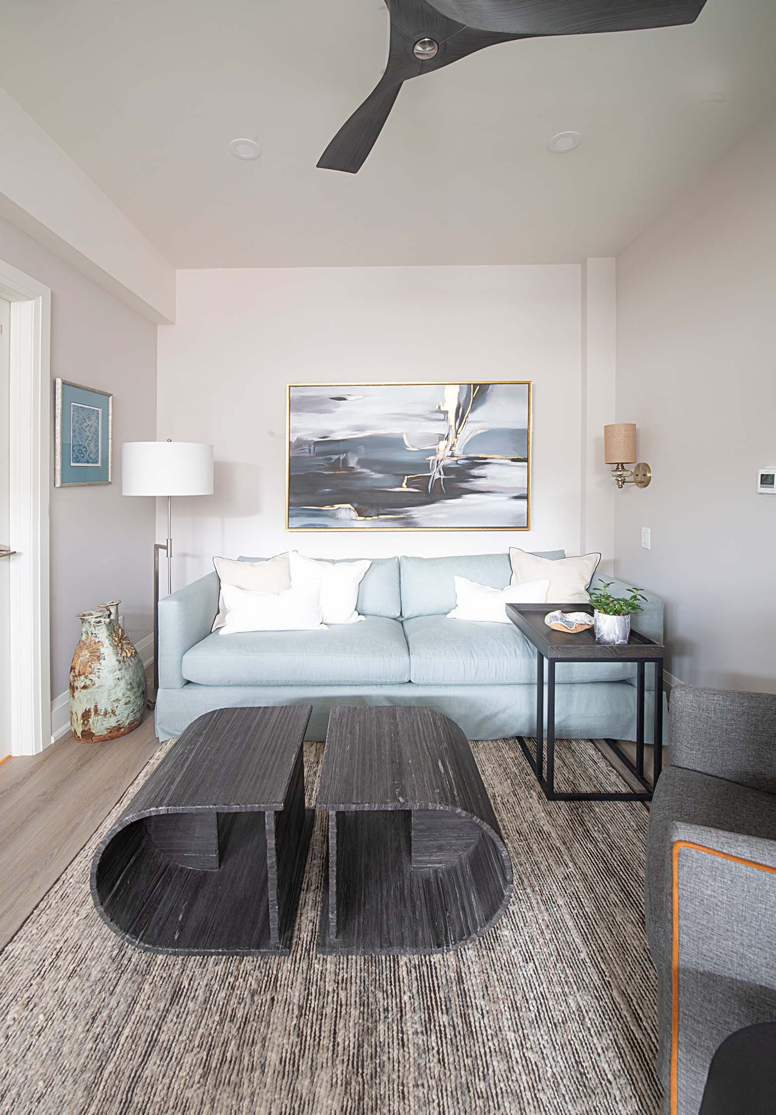 Contemporary living room with light blue sofa, abstract wall art, ceiling fan, and dark coffee tables on a textured rug.