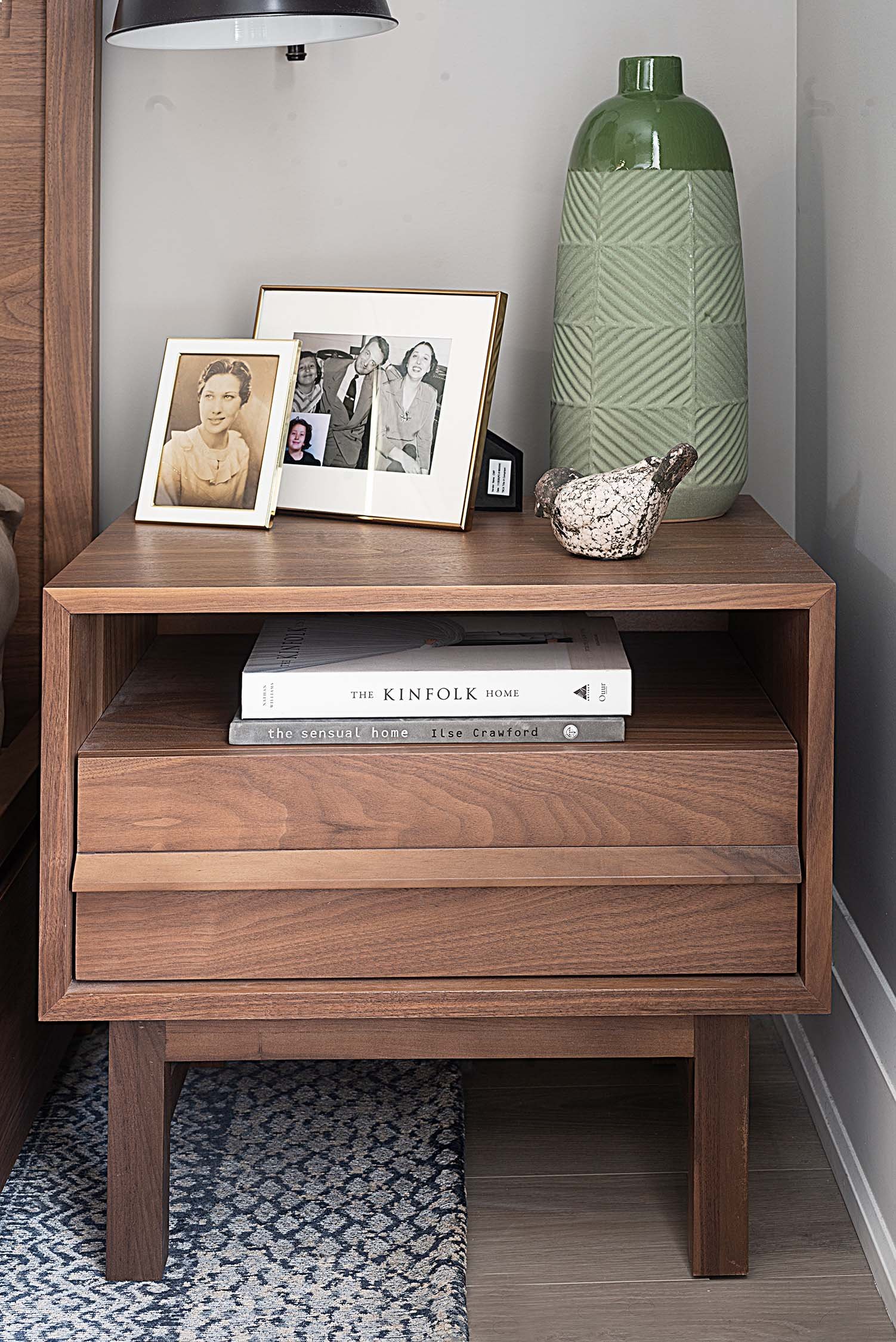 Wooden nightstand with framed photos, decorative vase, and books on a rug-covered floor.