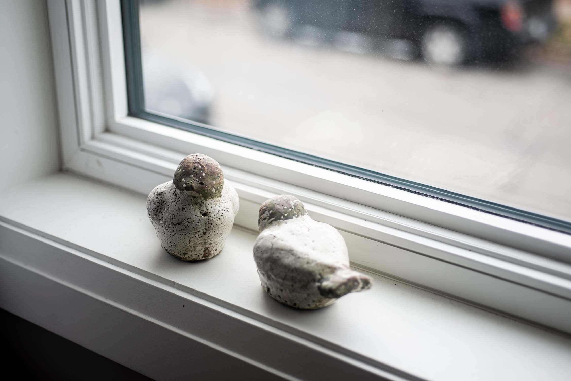 Two ceramic bird sculptures on a windowsill, next to a window overlooking a blurred street scene.