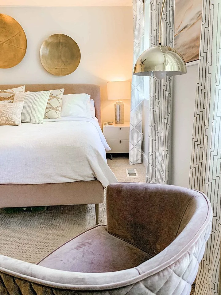 A bedroom with a beige bed, decorative pillows, a white nightstand with a lamp, a gold and silver round wall art, a metallic pendant light, and patterned curtains.