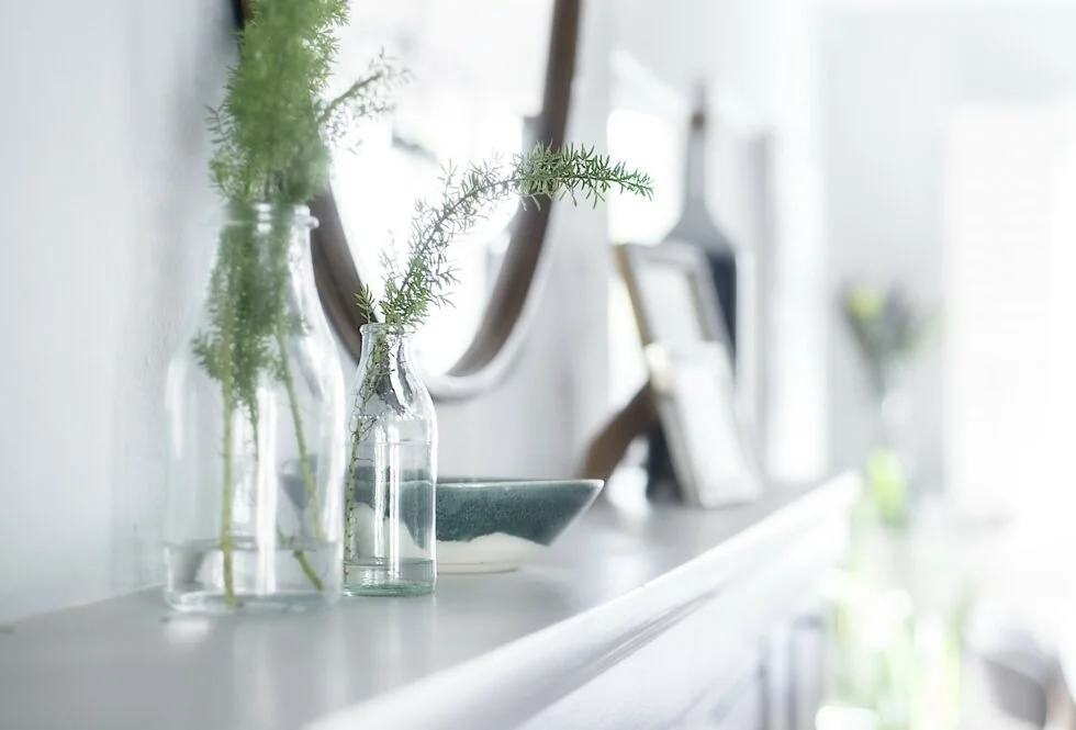 White mantle place with round mirror above. Decorative glass bottles, bowl and artwork sit on top of mantle place ledge.