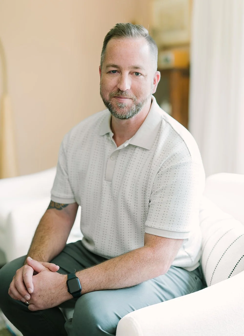 Man sitting on a white chair, wearing a light-colored polo shirt and a smartwatch, in a softly lit room.